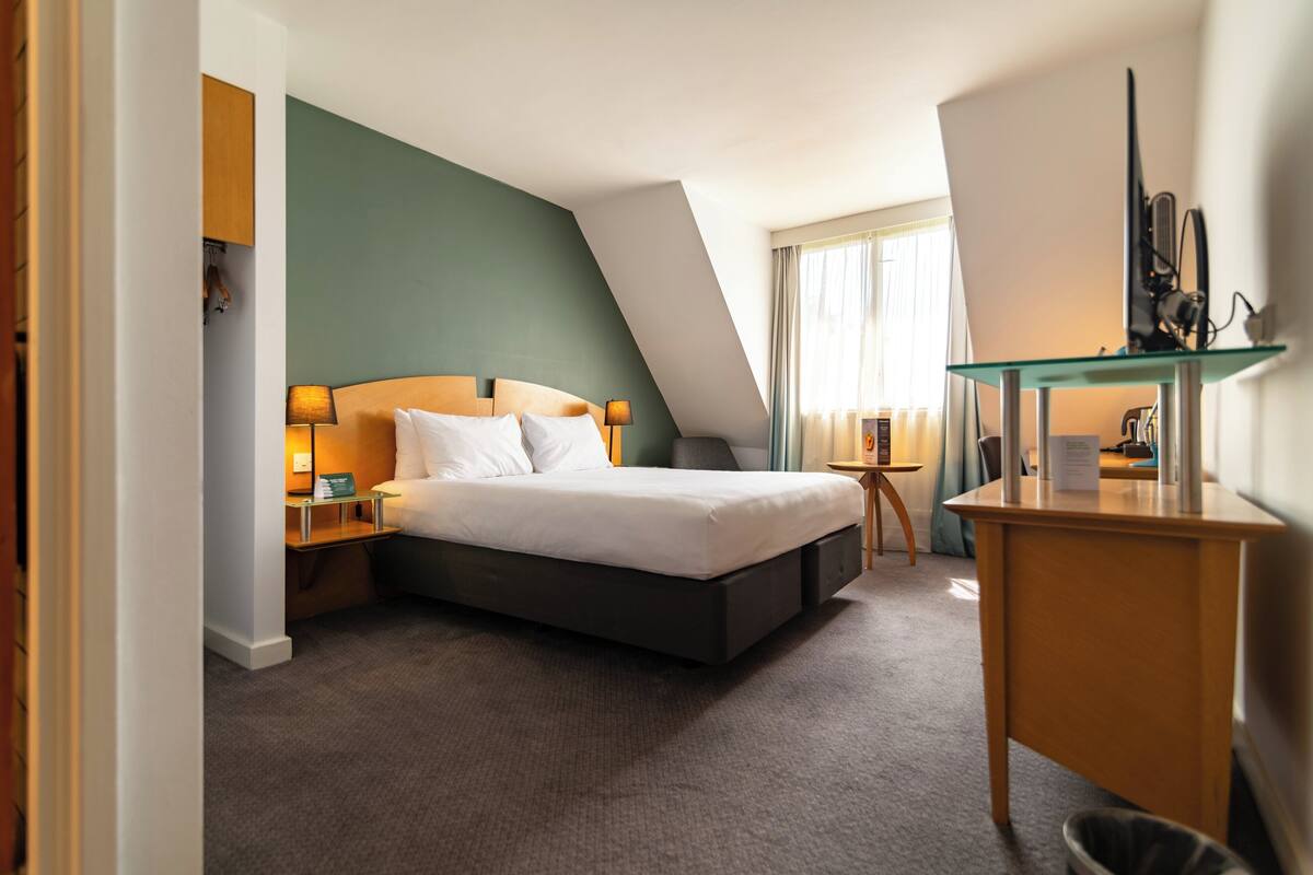 A modern hotel room with a large bed, a desk, and a TV on a glass shelf.