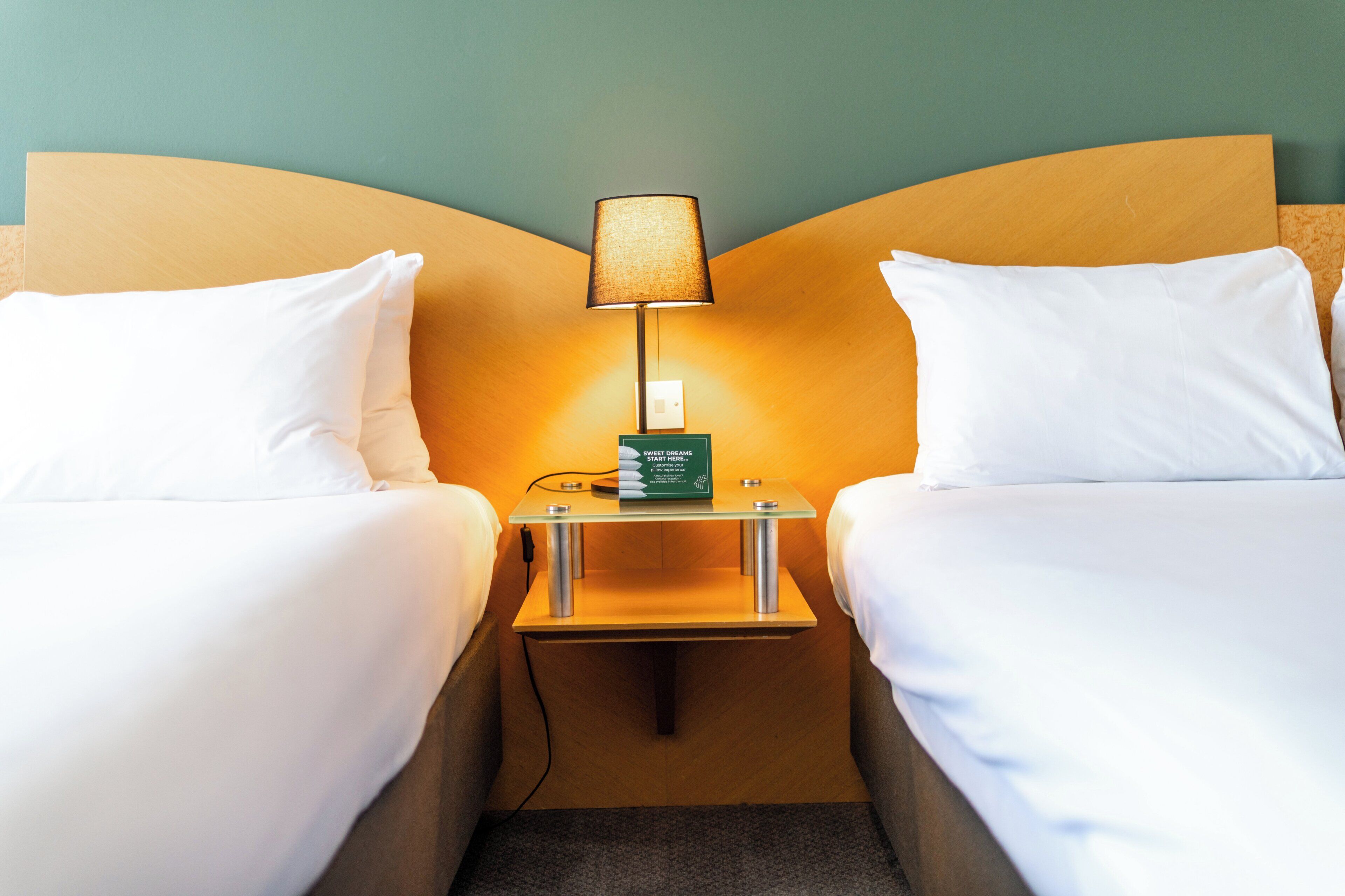Una habitación de hotel con dos camas individuales, una mesita de noche con una lámpara y una caja verde sobre la mesita.