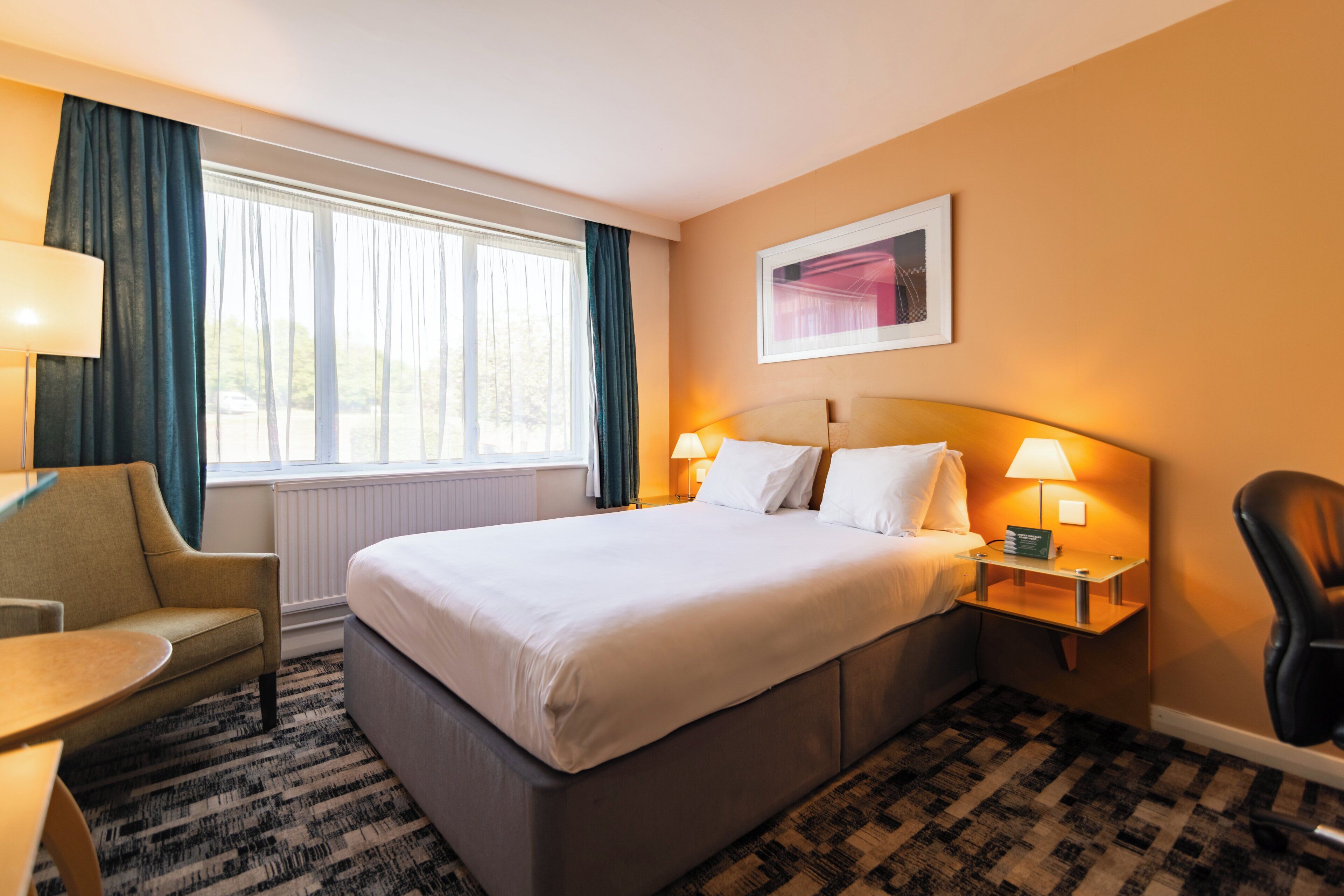 Una camera d'albergo con un letto grande, due comodini con lampade, una sedia, una scrivania e una finestra con tende.