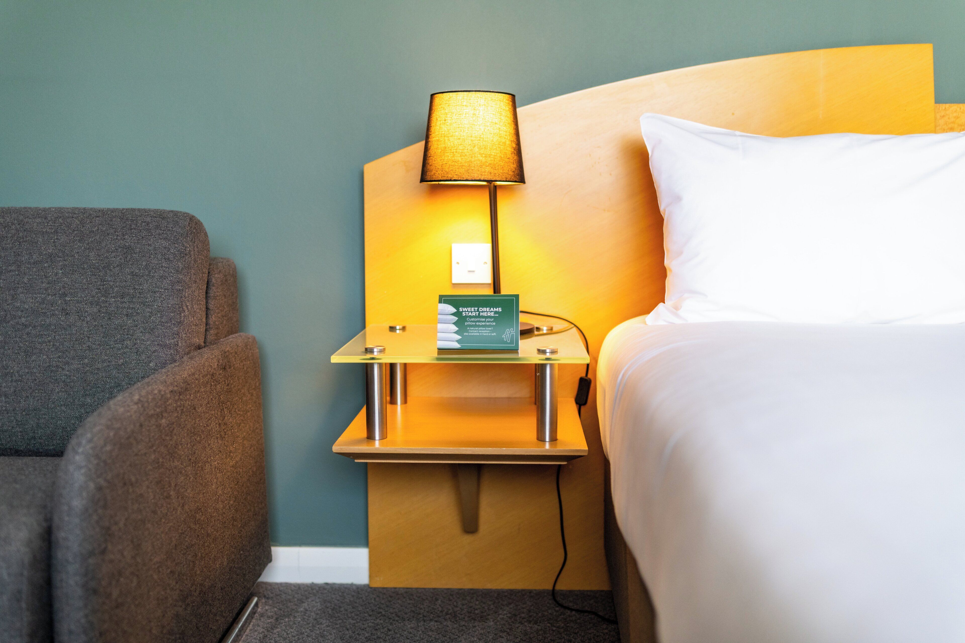 A hotel room with a bed, a nightstand with a lamp, a sign, and a grey armchair.