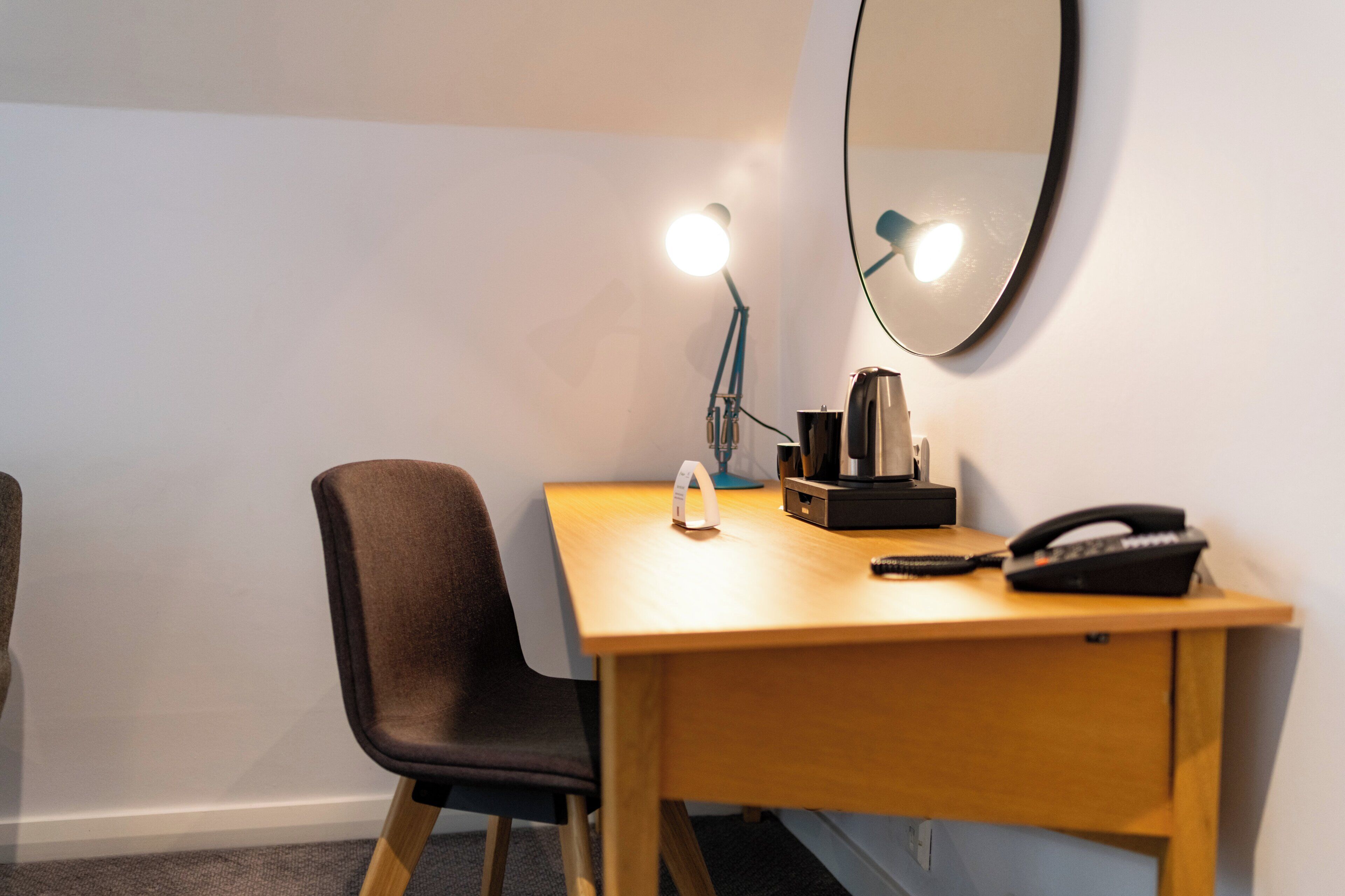 A hotel room with a wooden desk, a chair, a desk lamp, a telephone, and a mirror.