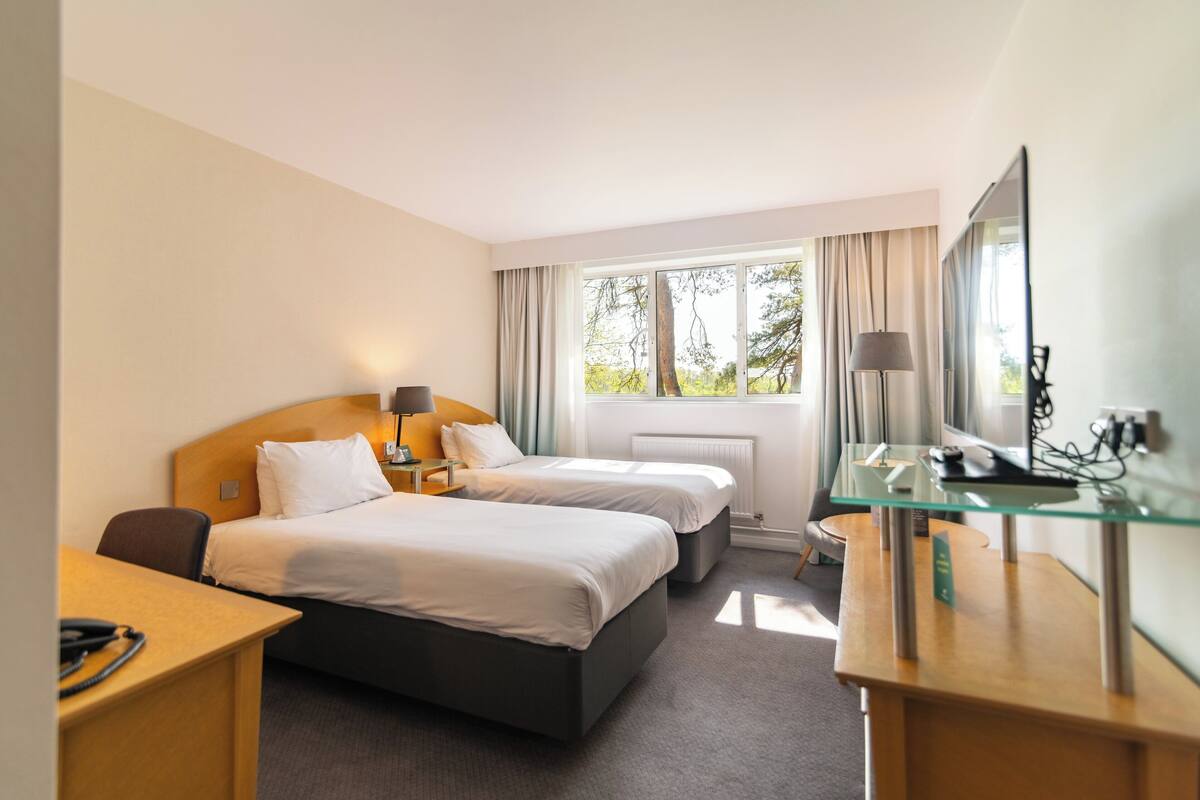 A hotel room with two beds, a desk, a chair, a TV, and a window with curtains.