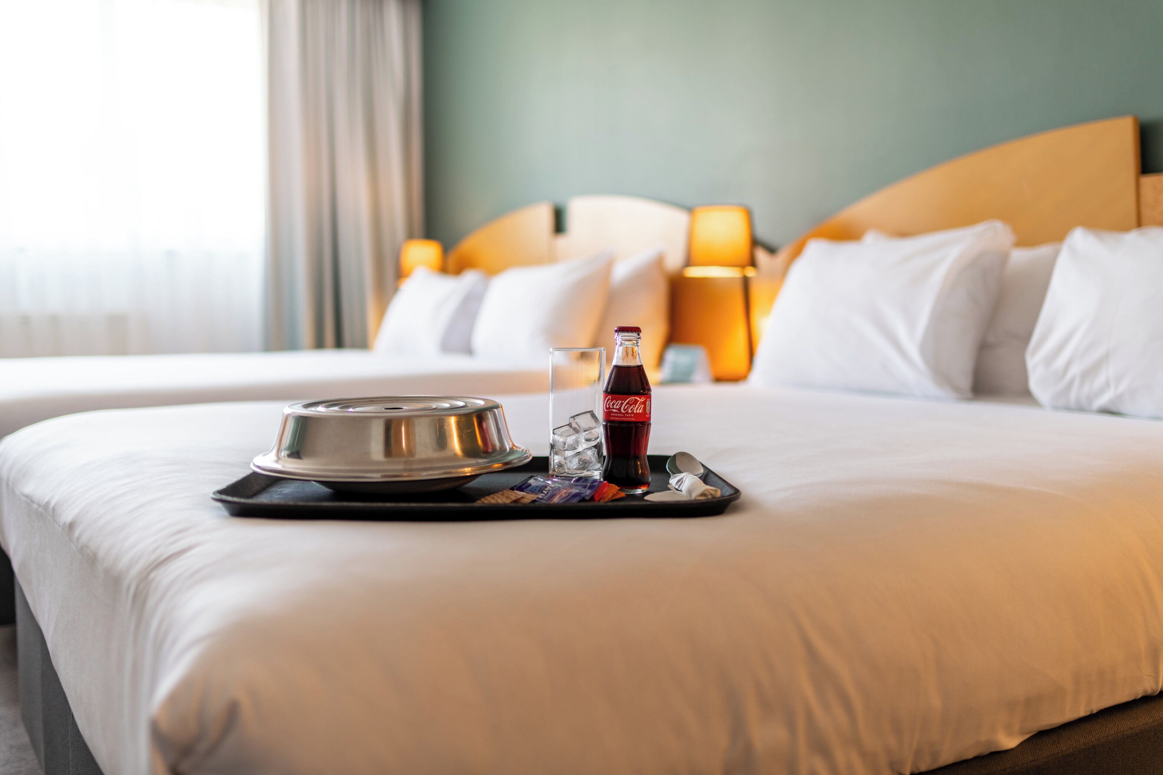 A neatly made bed with a tray containing a bottle of Coca-Cola, a glass, and a metal container, likely for breakfast.