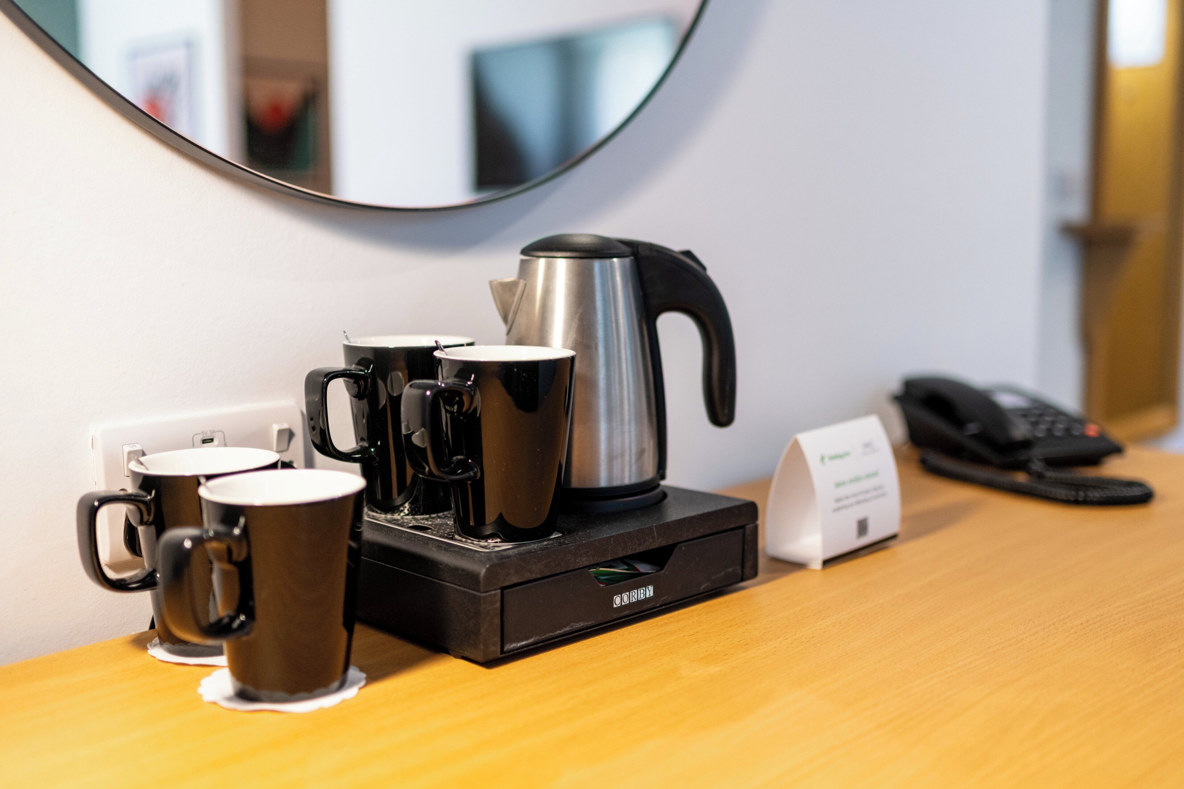 A coffee maker with two mugs on a wooden table, a kettle, a phone, and a small sign.