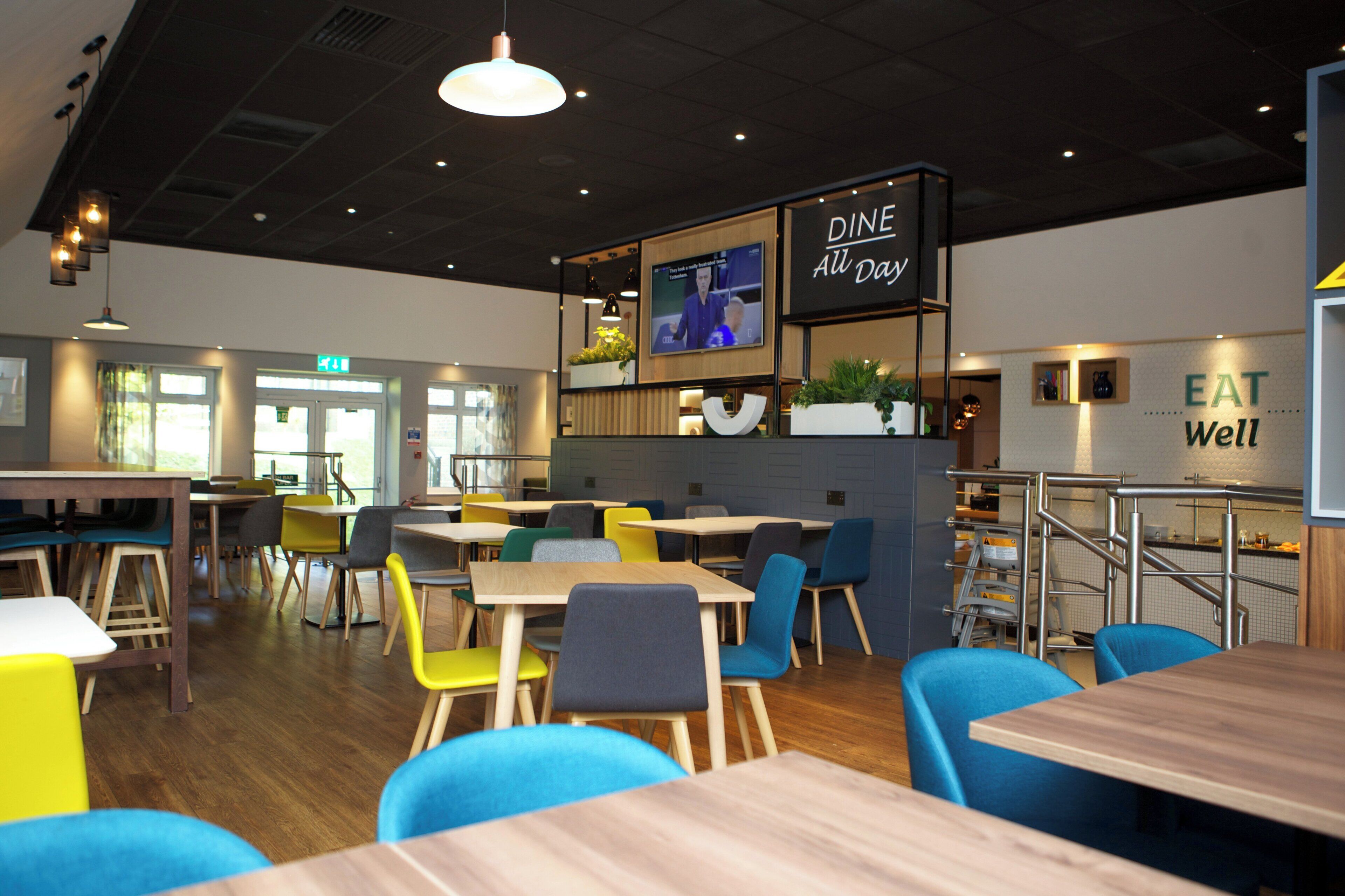 A modern restaurant interior with multiple dining tables and chairs, a television screen, and a sign that reads 'DINE All Day'.