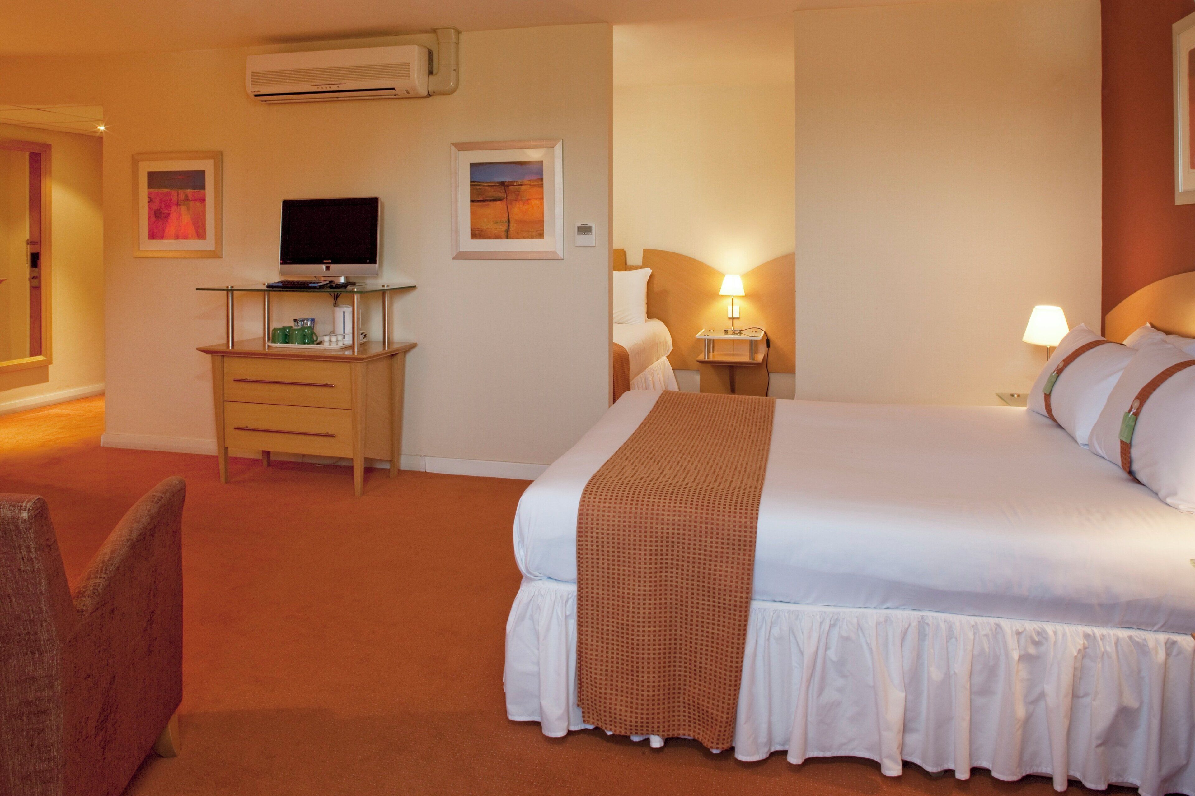 A hotel room with a bed, a desk with a computer, a TV, and a wall-mounted air conditioner.