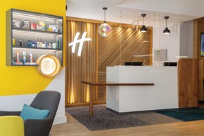 A reception desk with a modern design, a wooden slat wall with a 'H' logo, and a shelf with toiletries. - Holiday Inn Ashford North A20 by IHG (Ashford)