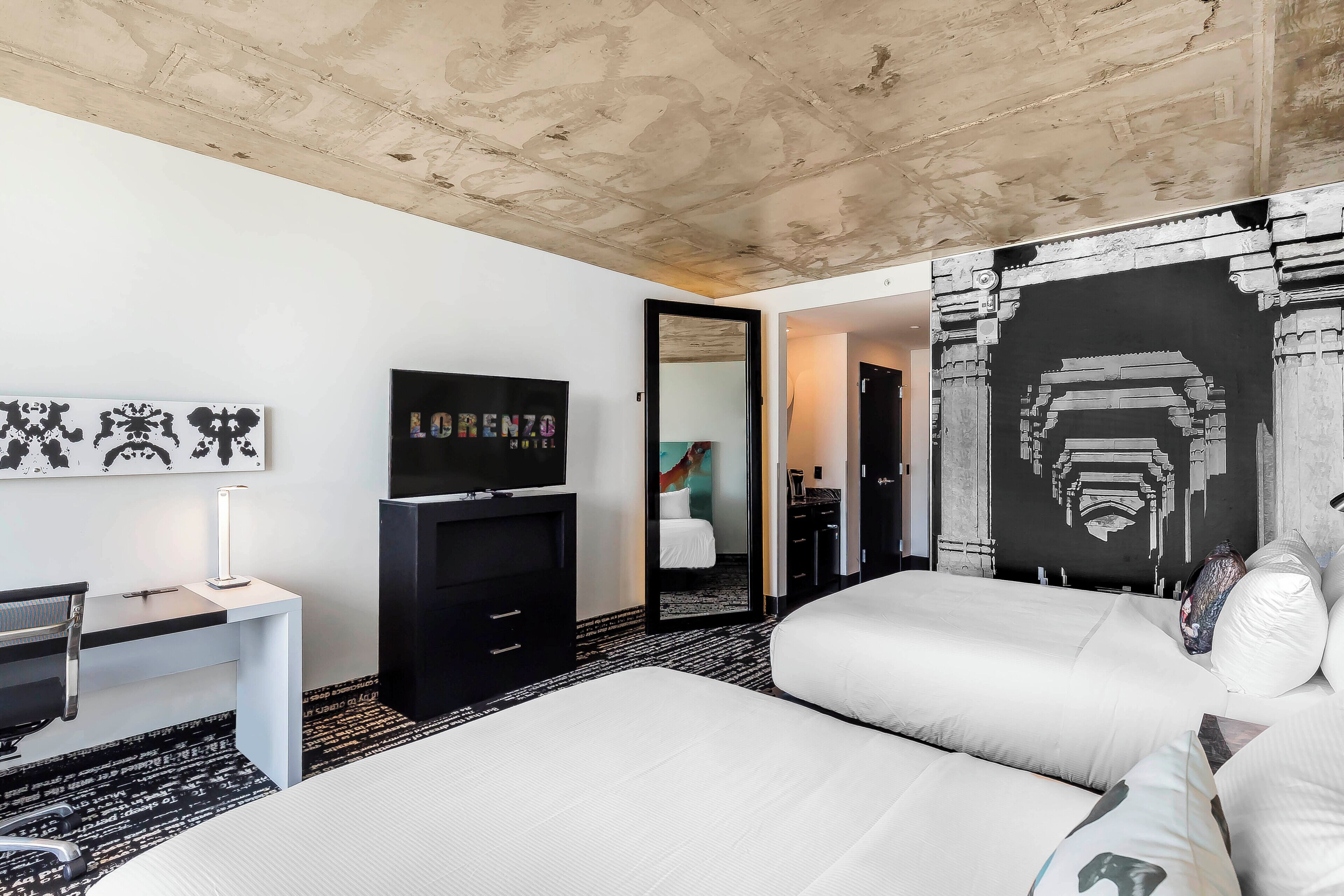 A hotel room with a bed, desk, TV, and artwork.