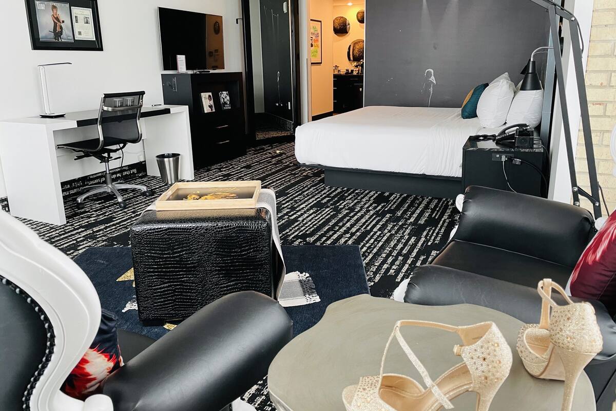 Hotellihuone, jossa on sänky, työpöytä, tuoli, sohva, lamppu ja pari korkeakorkoisia kenkiä sohvalla.