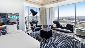 A modern hotel room with a large bed, two black leather sofas, a black floor lamp, and a view of the city through large windows. - Lorenzo Hotel Dallas, Tapestry Collection by Hilton (Dallas)