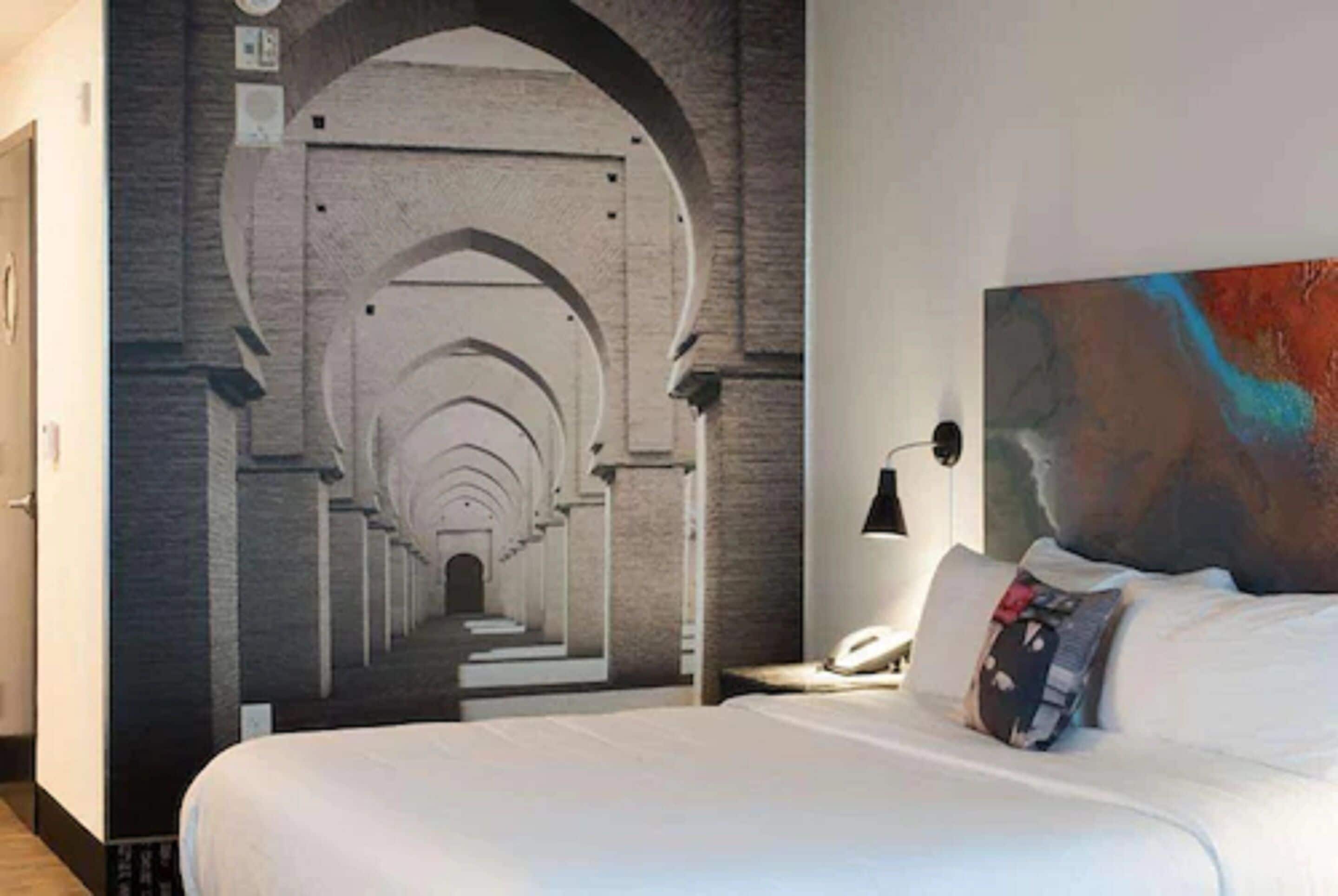 Una camera d'albergo con un letto grande, un comodino con una lampada e un affresco che raffigura un corridoio architettonico.