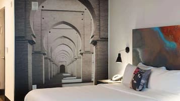 A hotel room with a large bed, a nightstand with a lamp, and a mural of an architectural corridor.