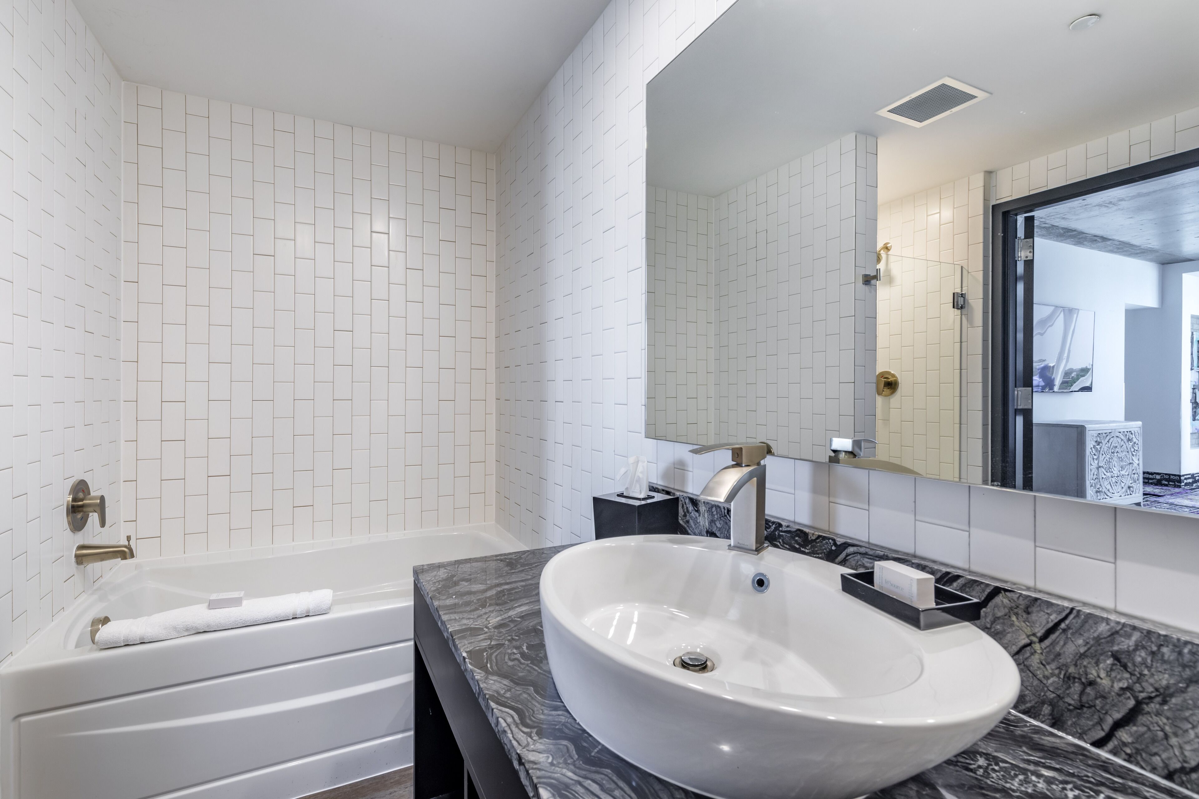 A modern bathroom with a white tiled wall, a white bathtub, a marble countertop, and a sink with a gold faucet.