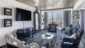 A modern hotel room with a large window, a black and white patterned carpet, a round glass coffee table, a dark sofa with decorative pillows, a black ornate dresser, and a mounted television. - Lorenzo Hotel Dallas, Tapestry Collection by Hilton (Dallas)