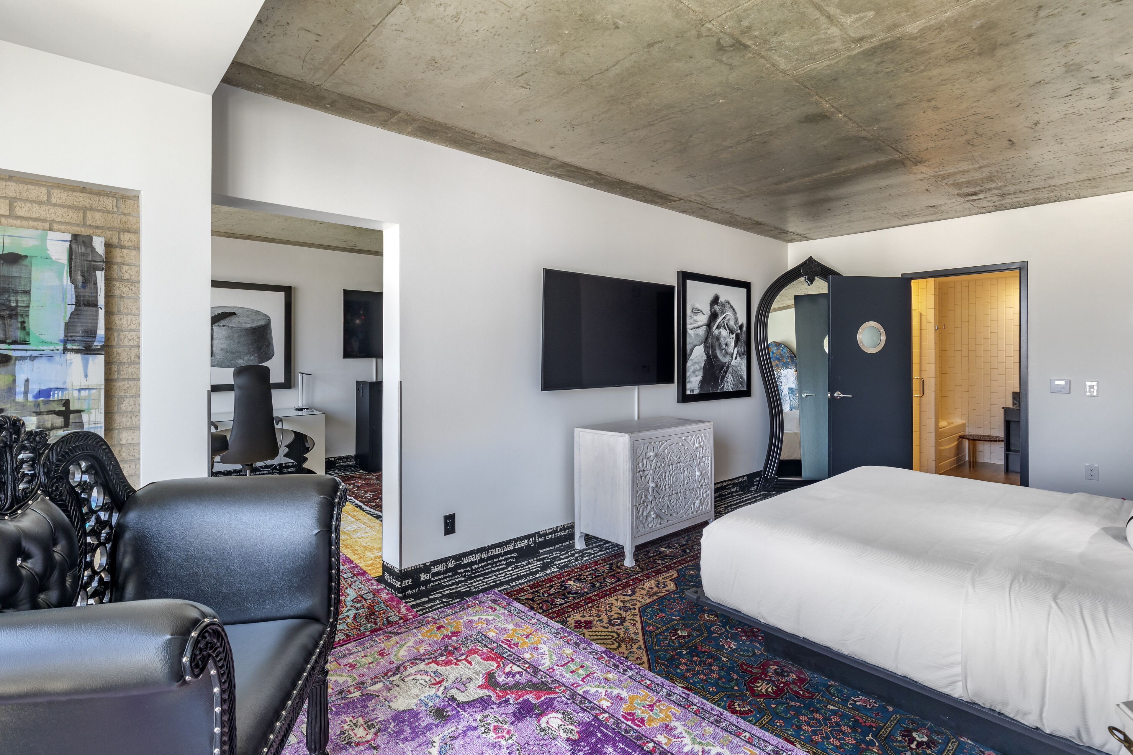 A modern hotel room with a large bed, a black leather chair, a flat-screen TV, and a decorative mirror.