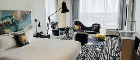 Ett modernt hotellrum med en stor säng, en svartvit fåtölj, ett skrivbord med en dator och ett stort fönster med gardiner.
