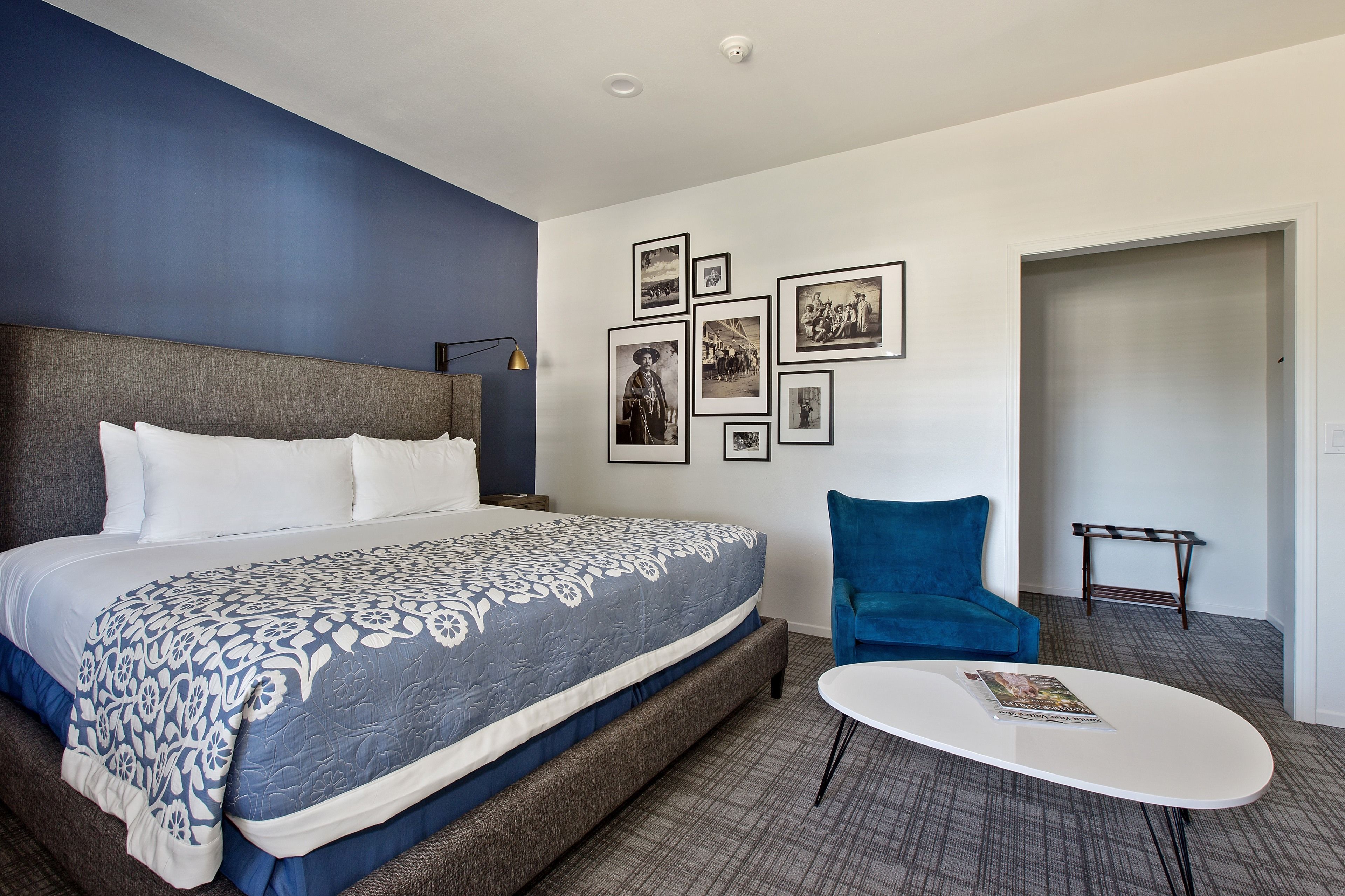 A hotel room with a large bed, a blue chair, a small table, and a wall with framed pictures.