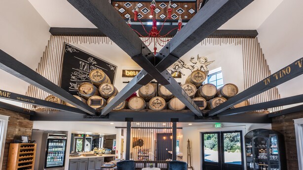 A bar with a high ceiling, exposed beams, and a chandelier. There are two blue chairs and a small table in the center.
