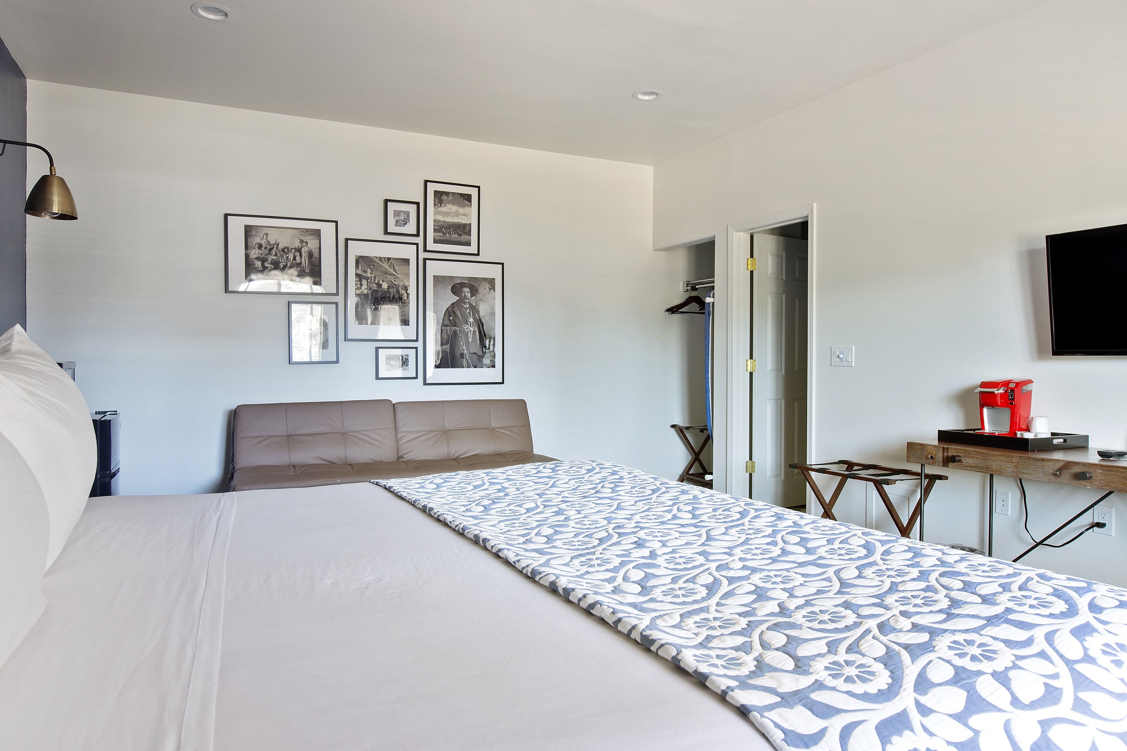 A bedroom with a large bed, a desk with a coffee maker, and several framed pictures on the wall.
