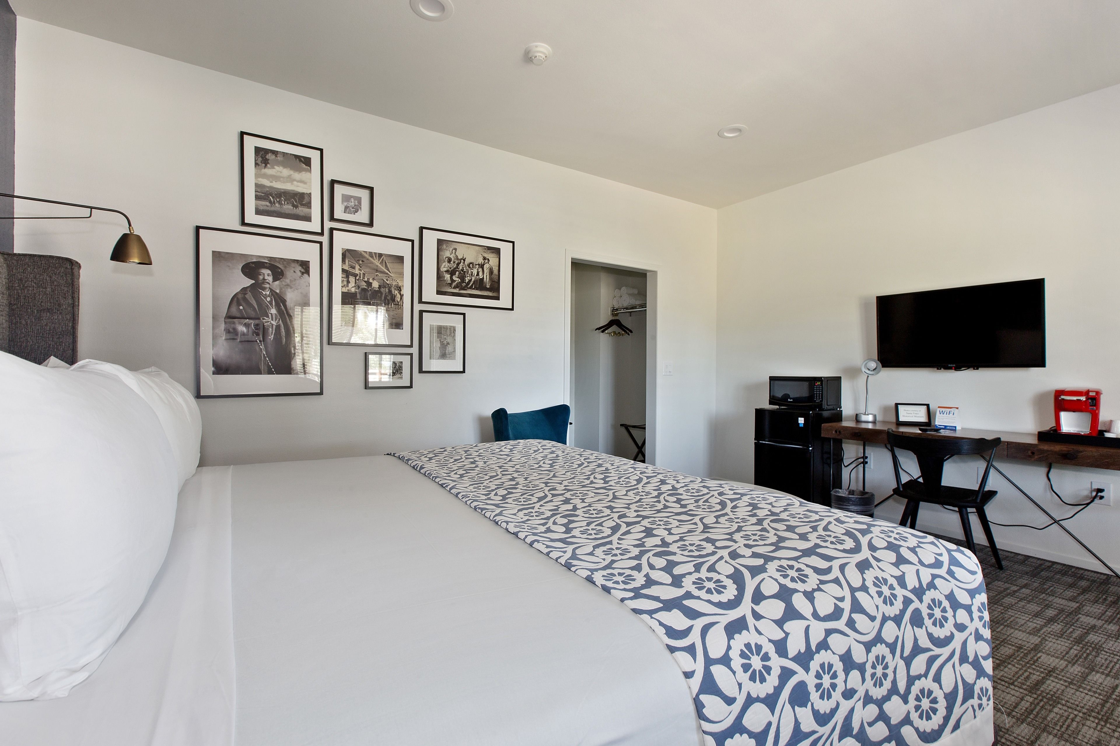 Una habitación de hotel con una cama, un escritorio con computadora, un televisor y varias fotos enmarcadas en la pared.