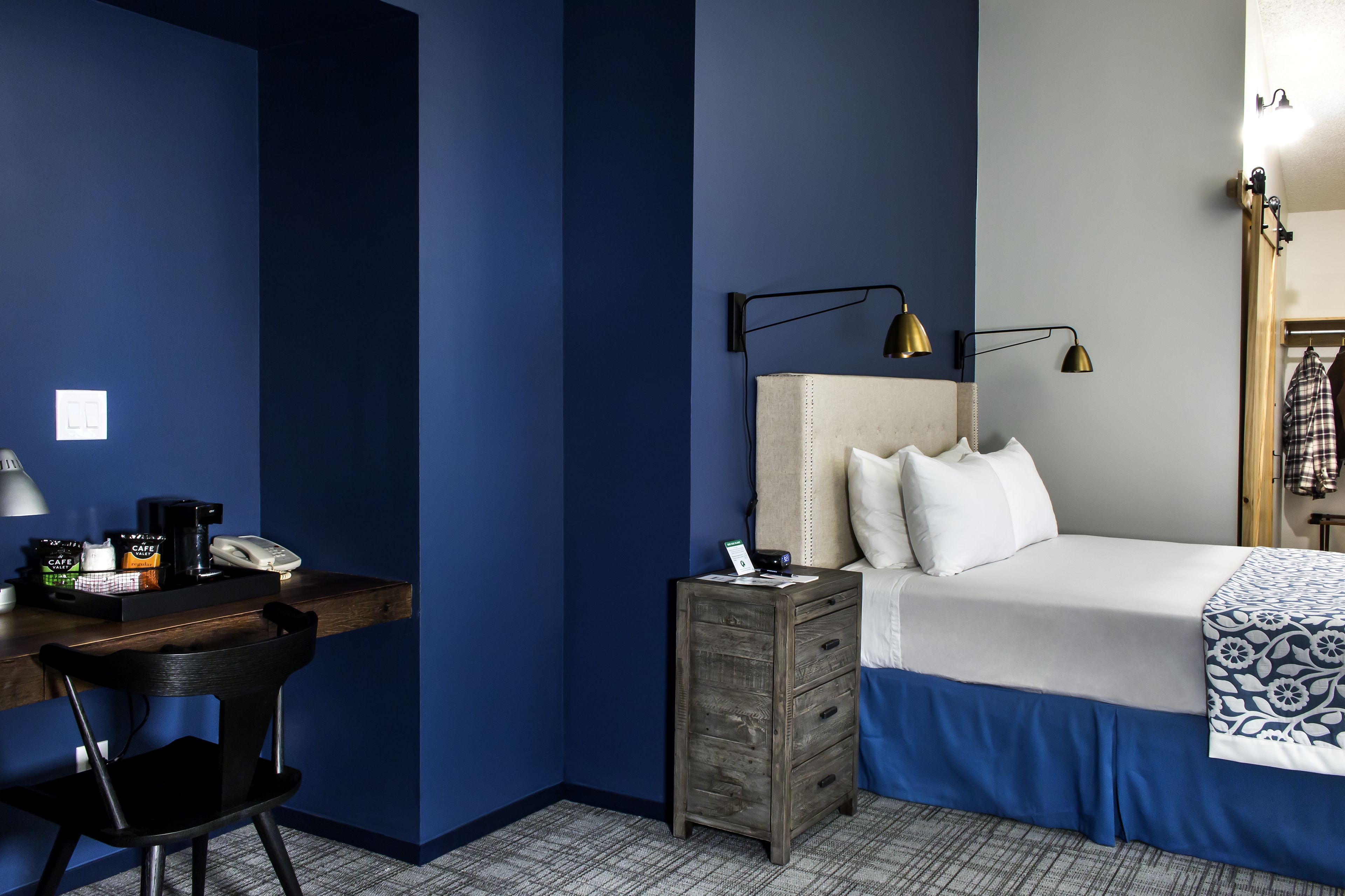 A hotel room with a bed, a desk with a chair, a nightstand, and a lamp.