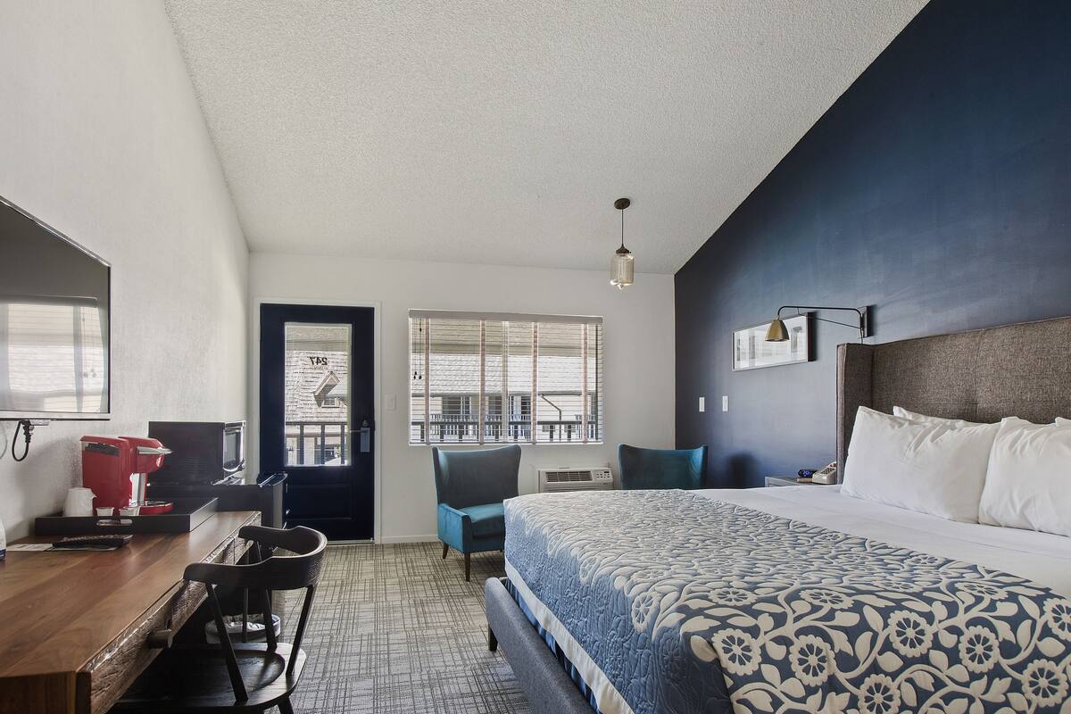 A hotel room with a bed, a desk with a coffee maker, a chair, and a window with blinds.