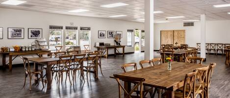 A spacious dining area with multiple wooden tables and chairs, large windows, and framed pictures on the walls.