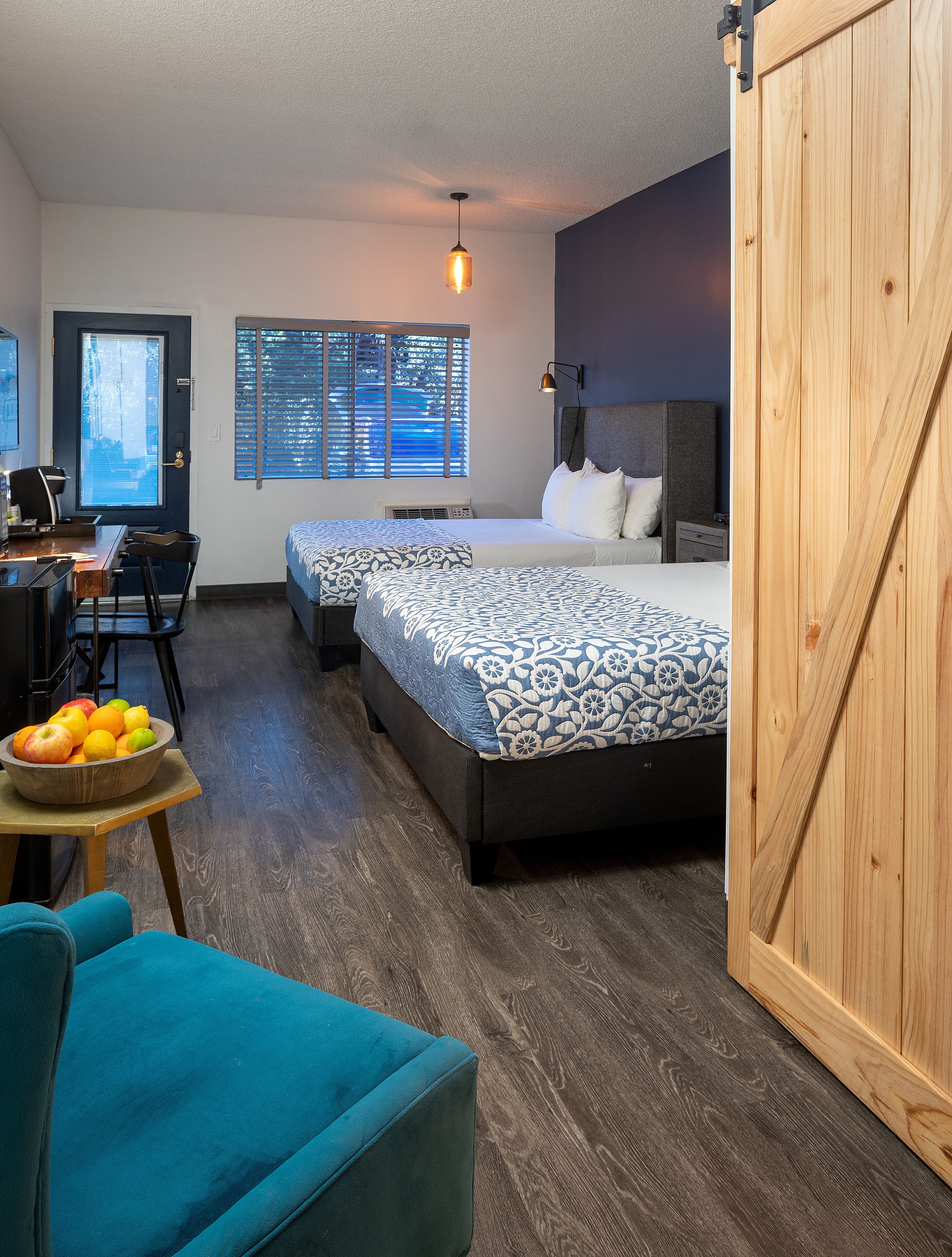 A hotel room with two beds, a desk, a chair, a wooden wardrobe, and a fruit bowl.