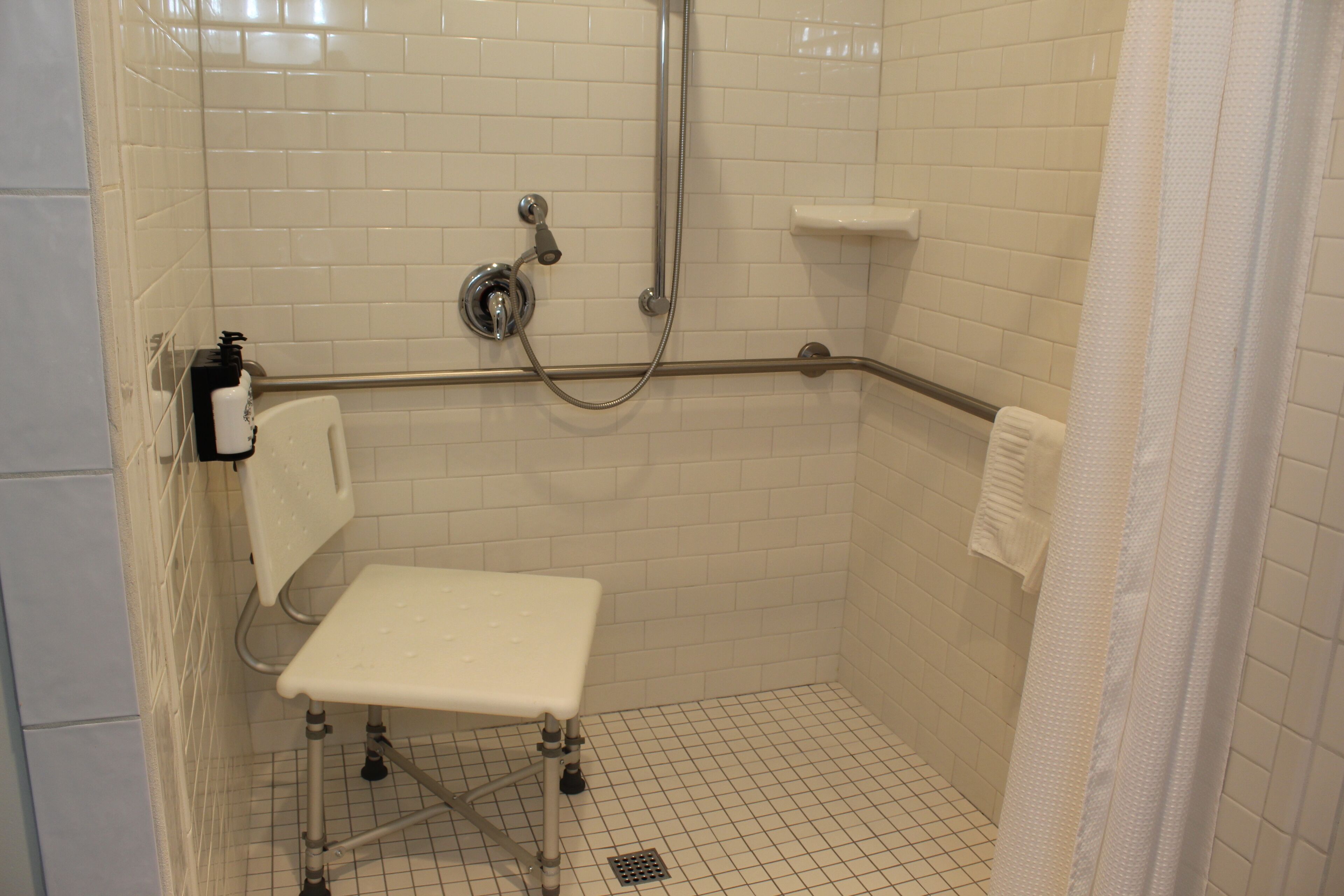 Une petite cabine de douche aux carreaux blancs, équipée d’une barre d’appui, d’un banc blanc et d’un pommeau de douche à main.