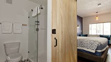 A bathroom with a toilet, shower, and glass door. There is a bed with a patterned bedspread and a hanging light fixture.