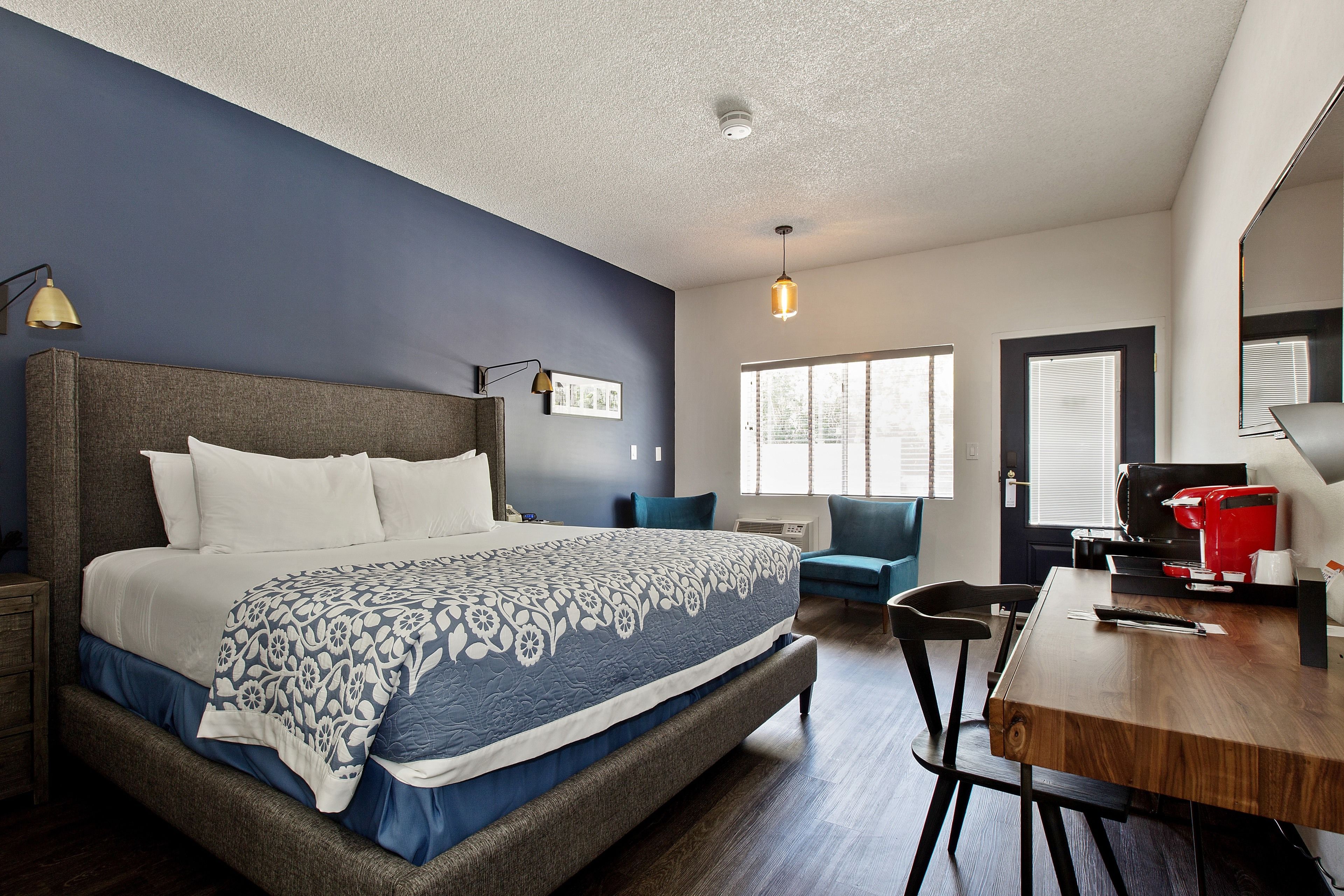 Une chambre avec un grand lit, un bureau et une chaise. Il y a une fenêtre avec des stores, une porte et un luminaire au plafond.