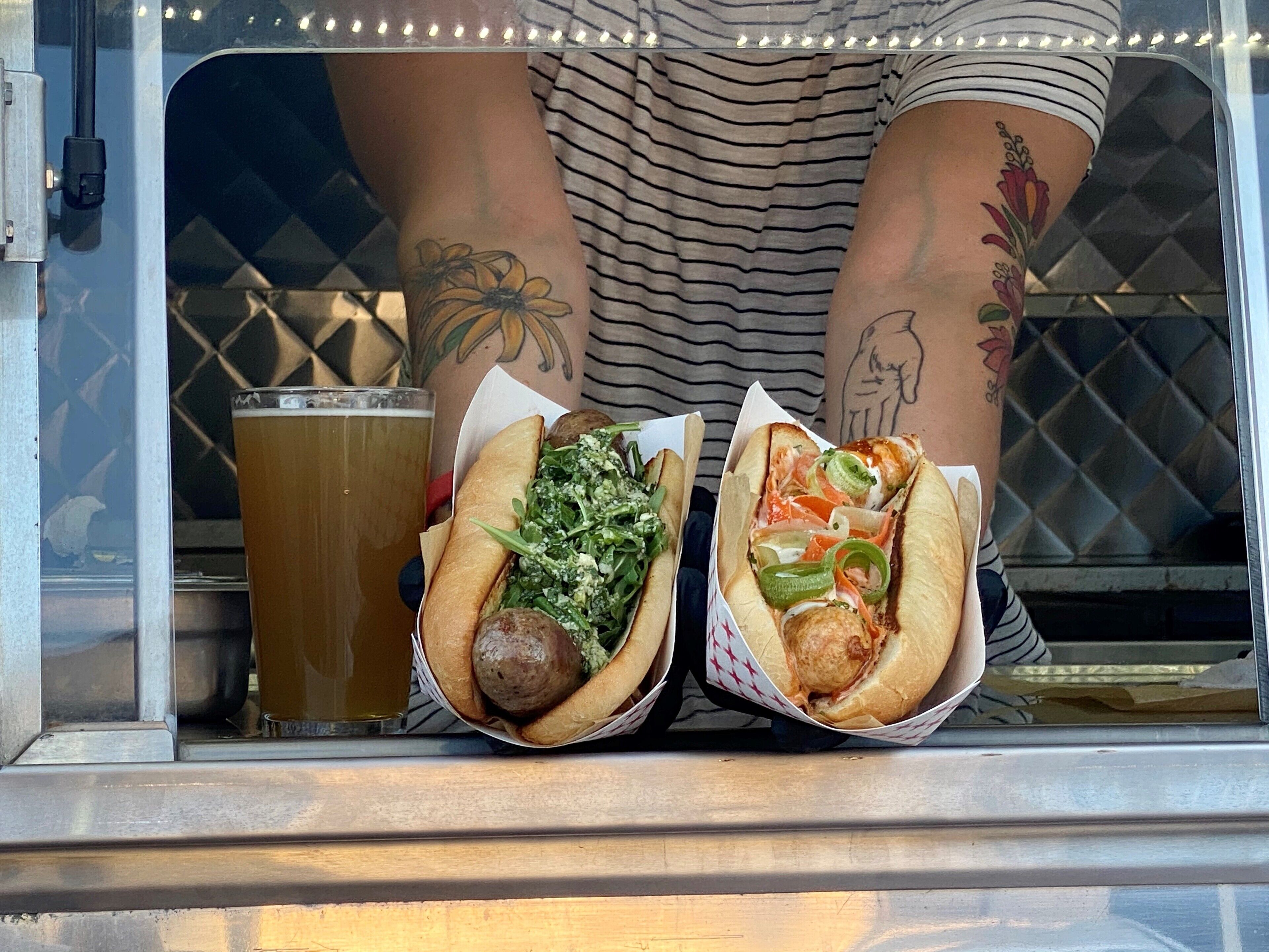 Henkilö, jolla on kädessään kaksi kuumaa hot dog -leipää, joissa on erilaisia täytteitä, sekä lasi olutta.