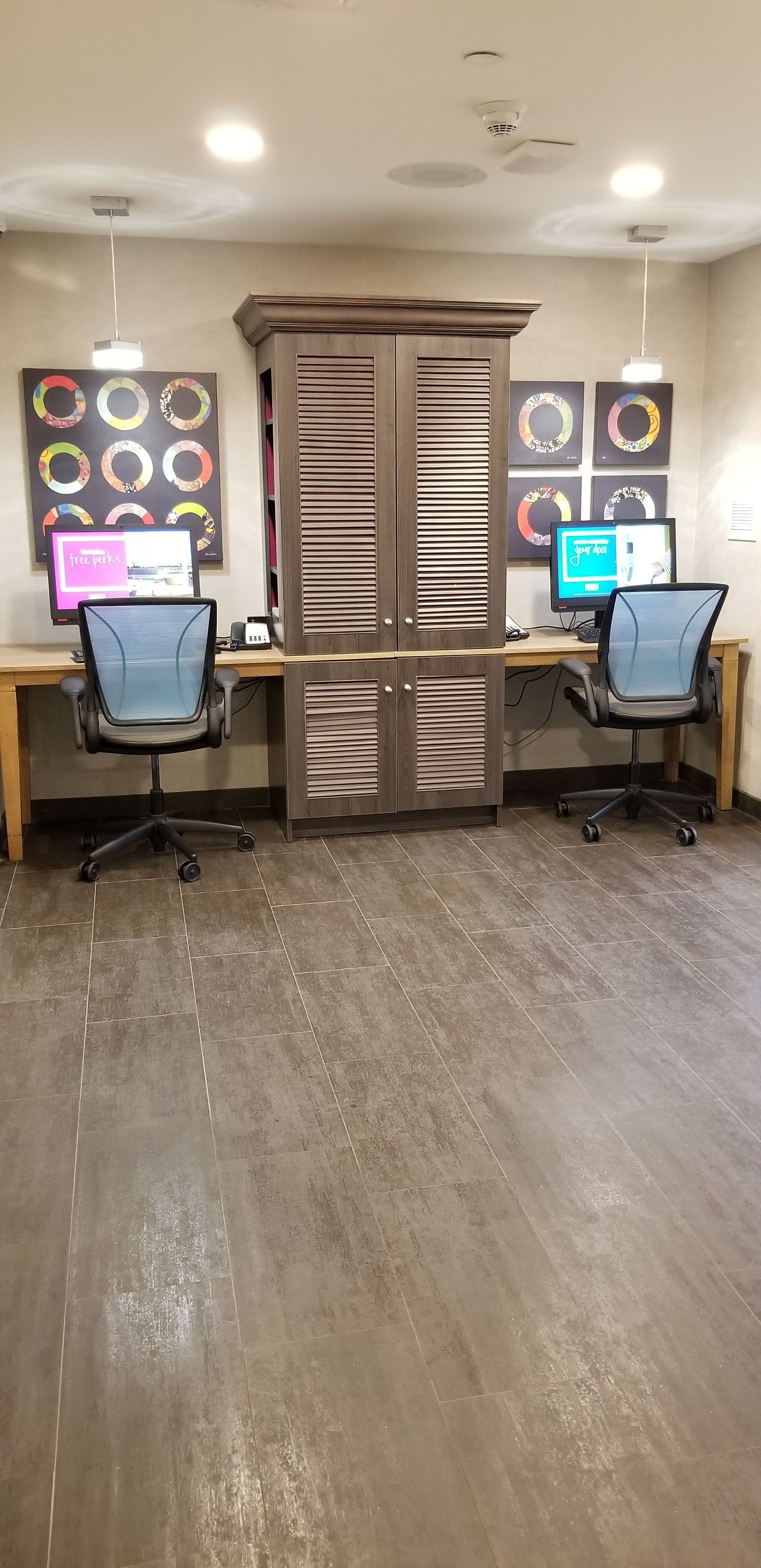 Un espace de bureau moderne comprenant deux bureaux, chacun équipé d’un ordinateur et d’une chaise. Des œuvres d’art décoratives ornent le mur au-dessus des bureaux, et un grand meuble est placé au centre de la pièce.