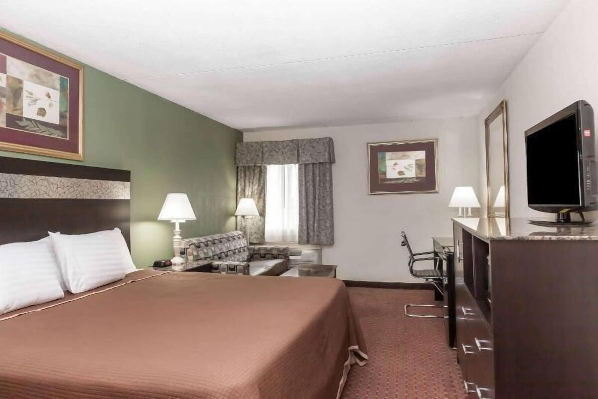 A hotel room with a bed, two bedside lamps, a television, a desk with a chair, and a window with curtains.
