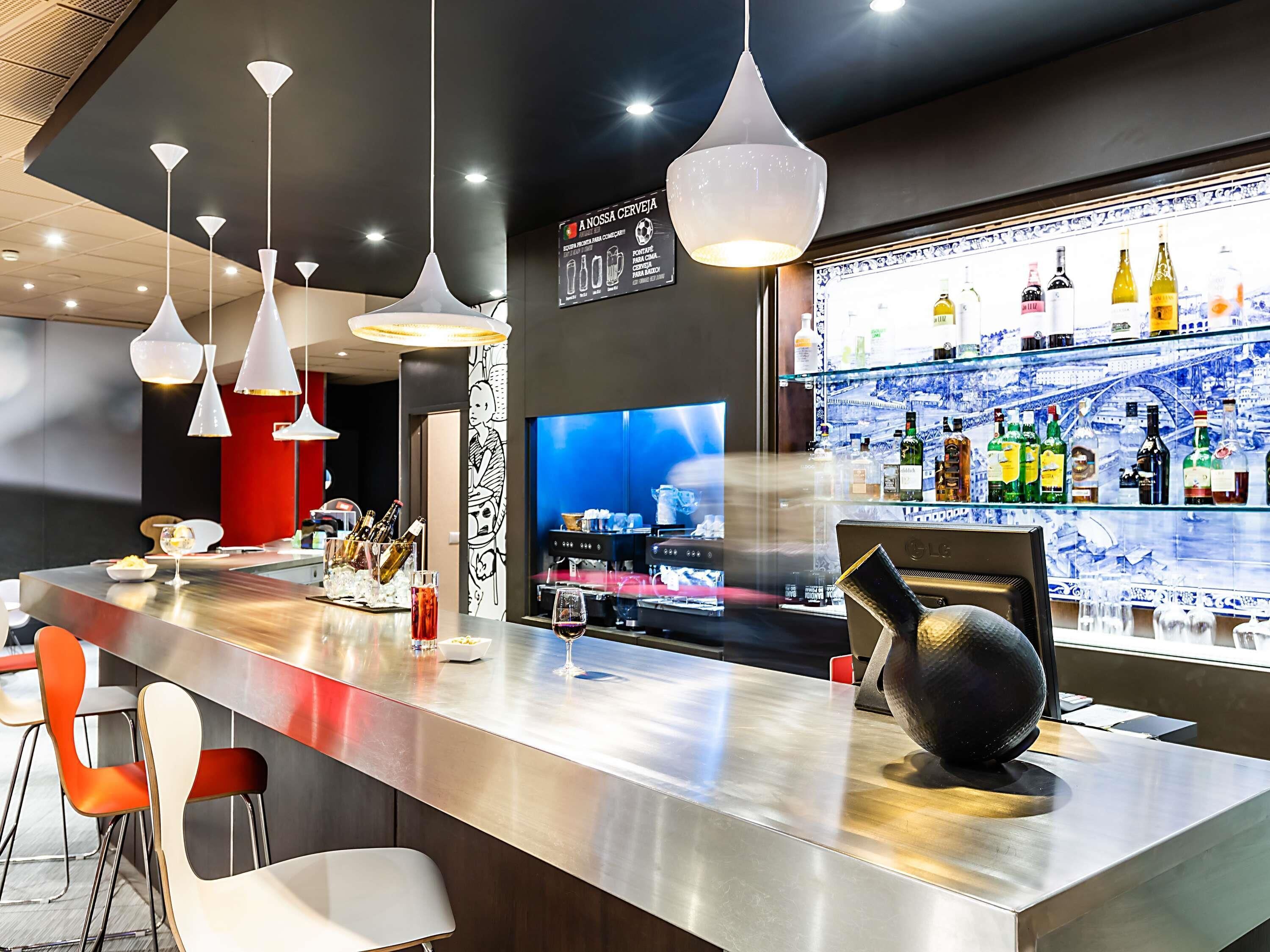 A modern bar area with a long counter, multiple hanging pendant lights, a television screen, and a variety of bottles on shelves.