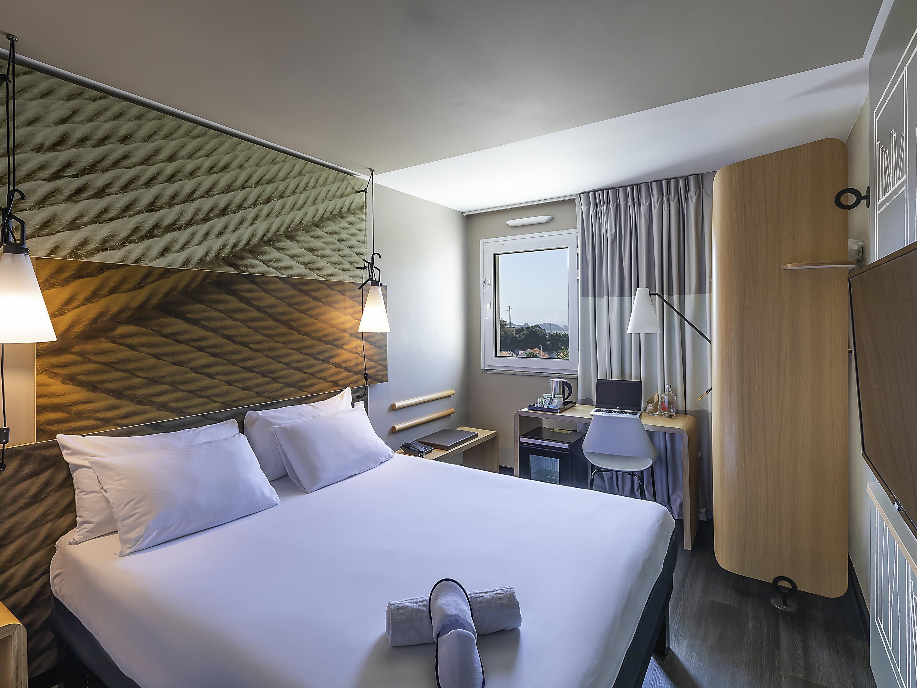A hotel room with a bed, a desk, a chair, a window with curtains, and a headboard with a textured design.