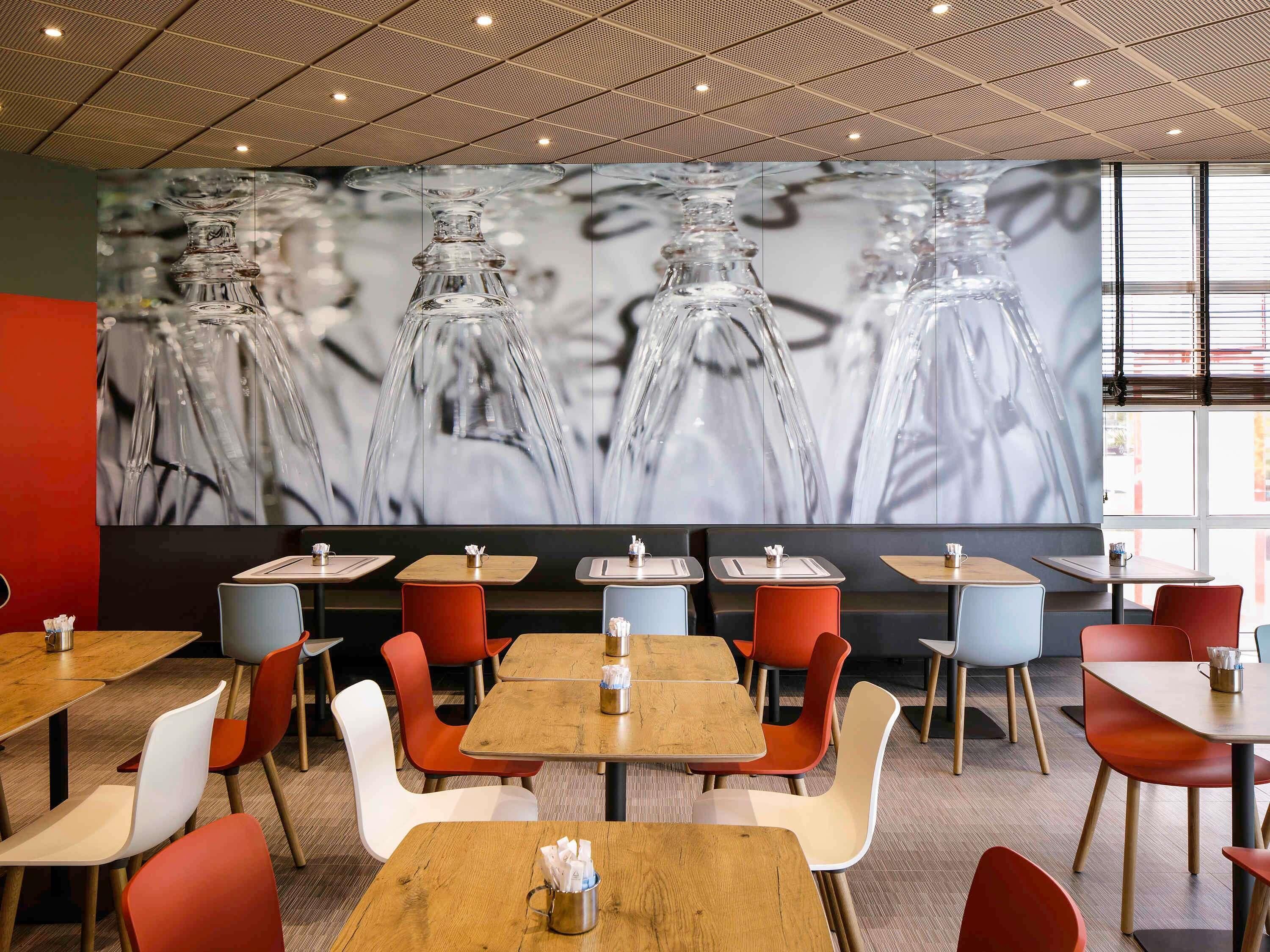 A modern restaurant interior with wooden tables and colorful chairs, a large mural of glass bottles, and a patterned ceiling.