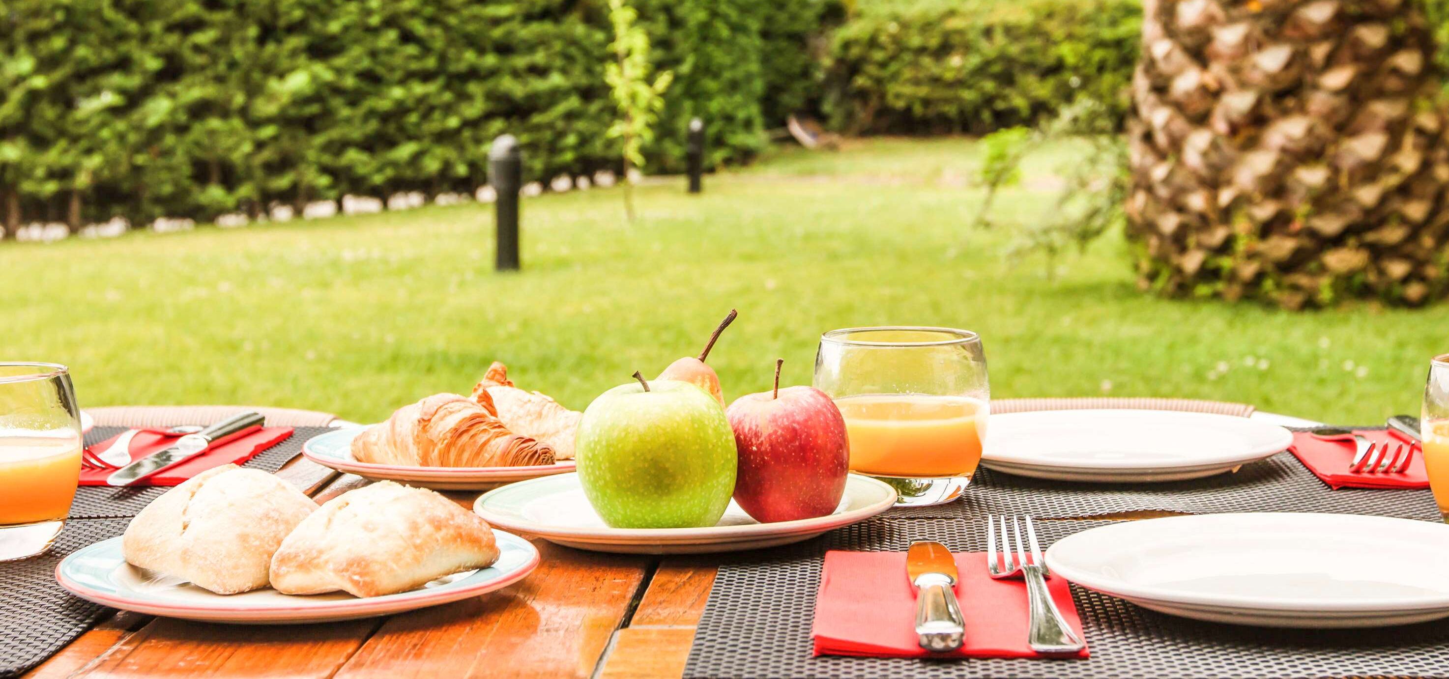 Un montaje para picnic con platos de comida, incluyendo cruasanes y manzanas, y vasos de jugo de naranja sobre una mesa de madera al aire libre.