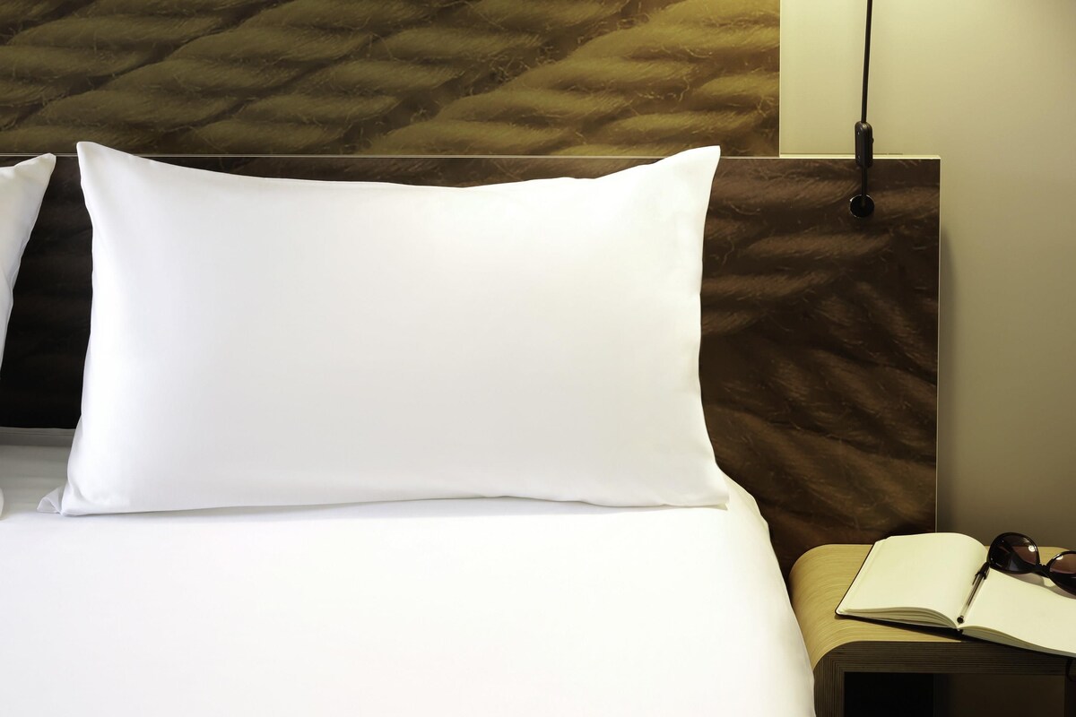 A neatly made bed with white bedding, a headboard with a textured pattern, and a bedside table with an open book and sunglasses.