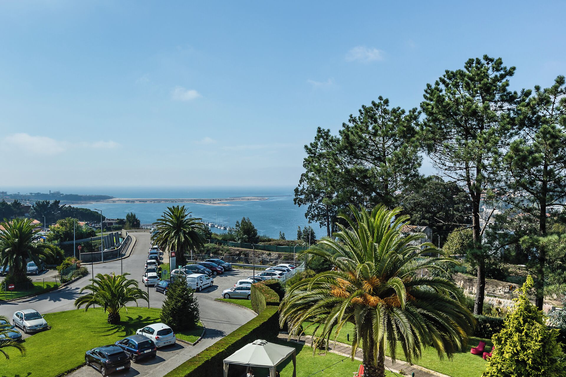 Un parking rempli de voitures, un petit bâtiment et une vue sur la mer et le paysage urbain lointain.