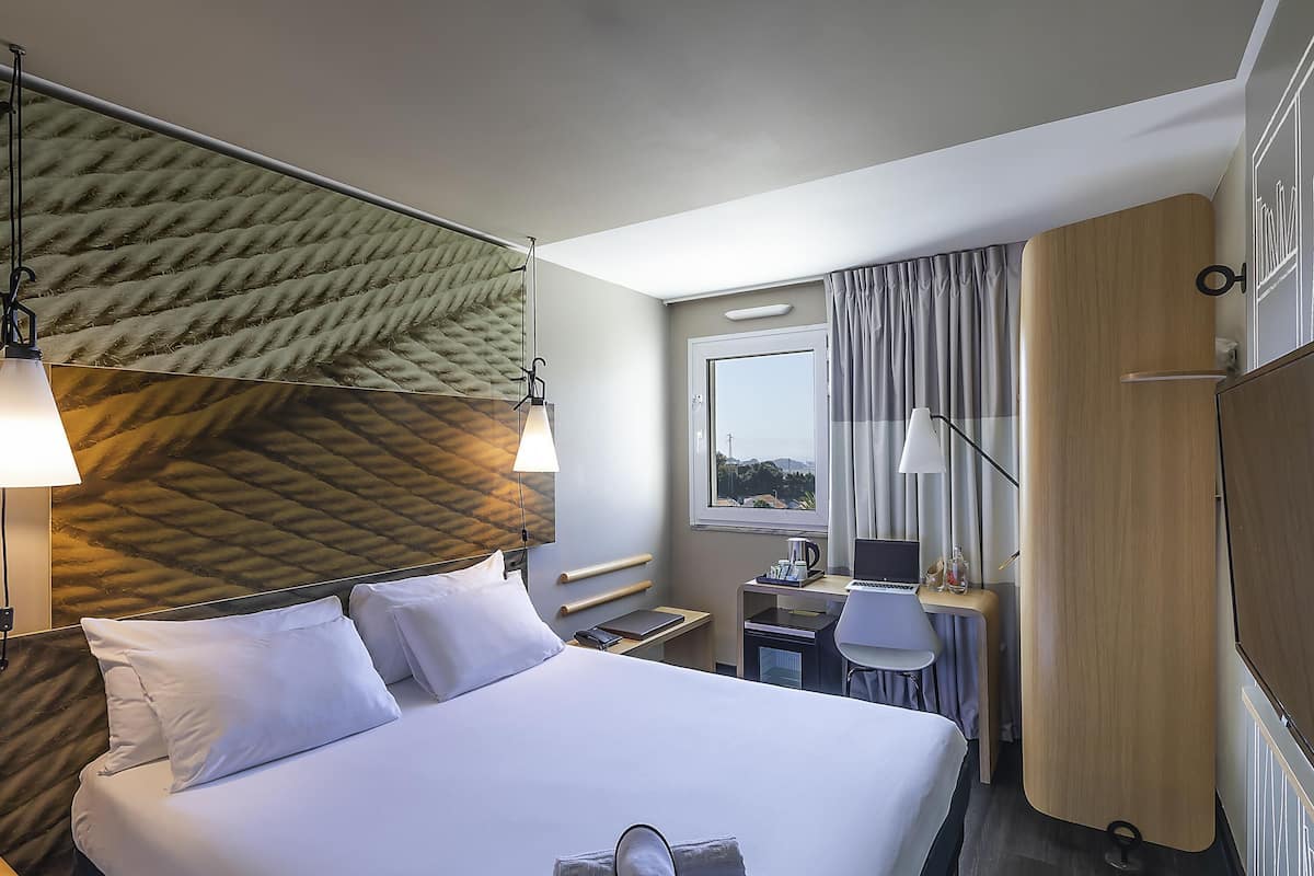 Ein Hotelzimmer mit Bett, Schreibtisch, Stuhl, einem Fenster mit Vorhängen und einem Kopfteil mit strukturierter Gestaltung.
