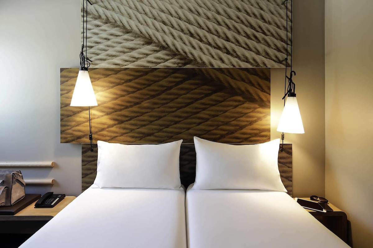 A double bed with white linens, two bedside lamps, a wooden headboard with a geometric pattern, a nightstand with a phone, and a wall-mounted mirror.