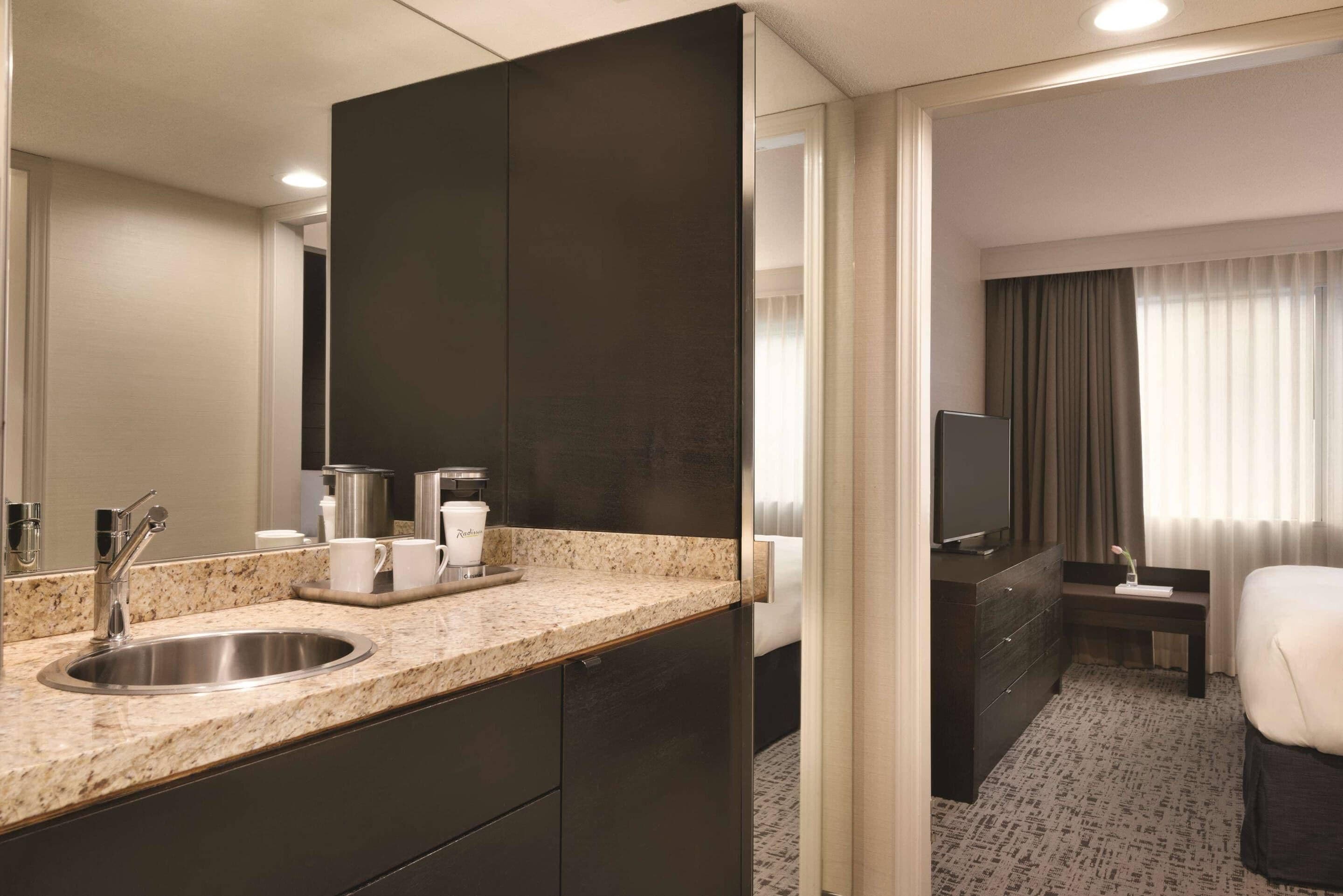 A modern hotel room with a granite countertop sink, dark cabinetry, a flat-screen TV, a bedside table, and a large mirror.