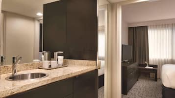 A modern hotel room with a granite countertop sink, dark cabinetry, a flat-screen TV, a bedside table, and a large mirror.
