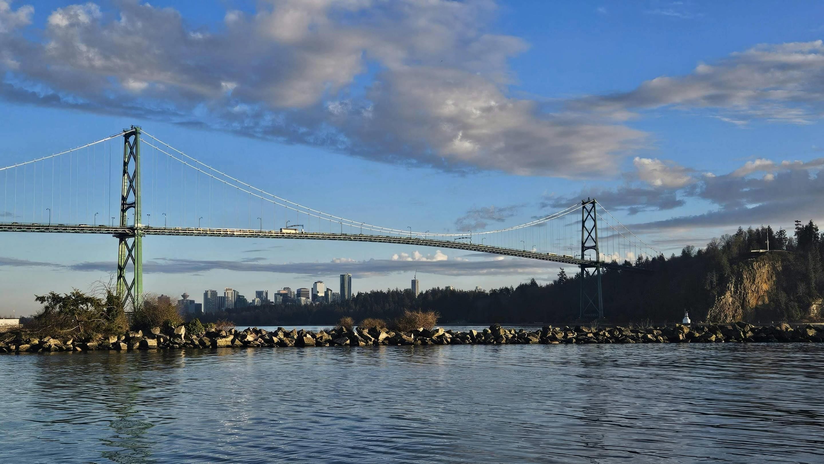 Un puente colgante sobre un cuerpo de agua, con el horizonte de una ciudad al fondo.