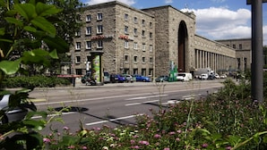 A large, historic building with a prominent archway, surrounded by a street with parked cars and greenery.