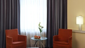 A hotel room with two orange armchairs, a small round table with a vase of flowers and fruit, and a large window with sheer curtains.