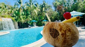 A coconut drink with a straw, a cherry, and a slice of fruit on a table by a pool with a waterfall and palm trees in the background.