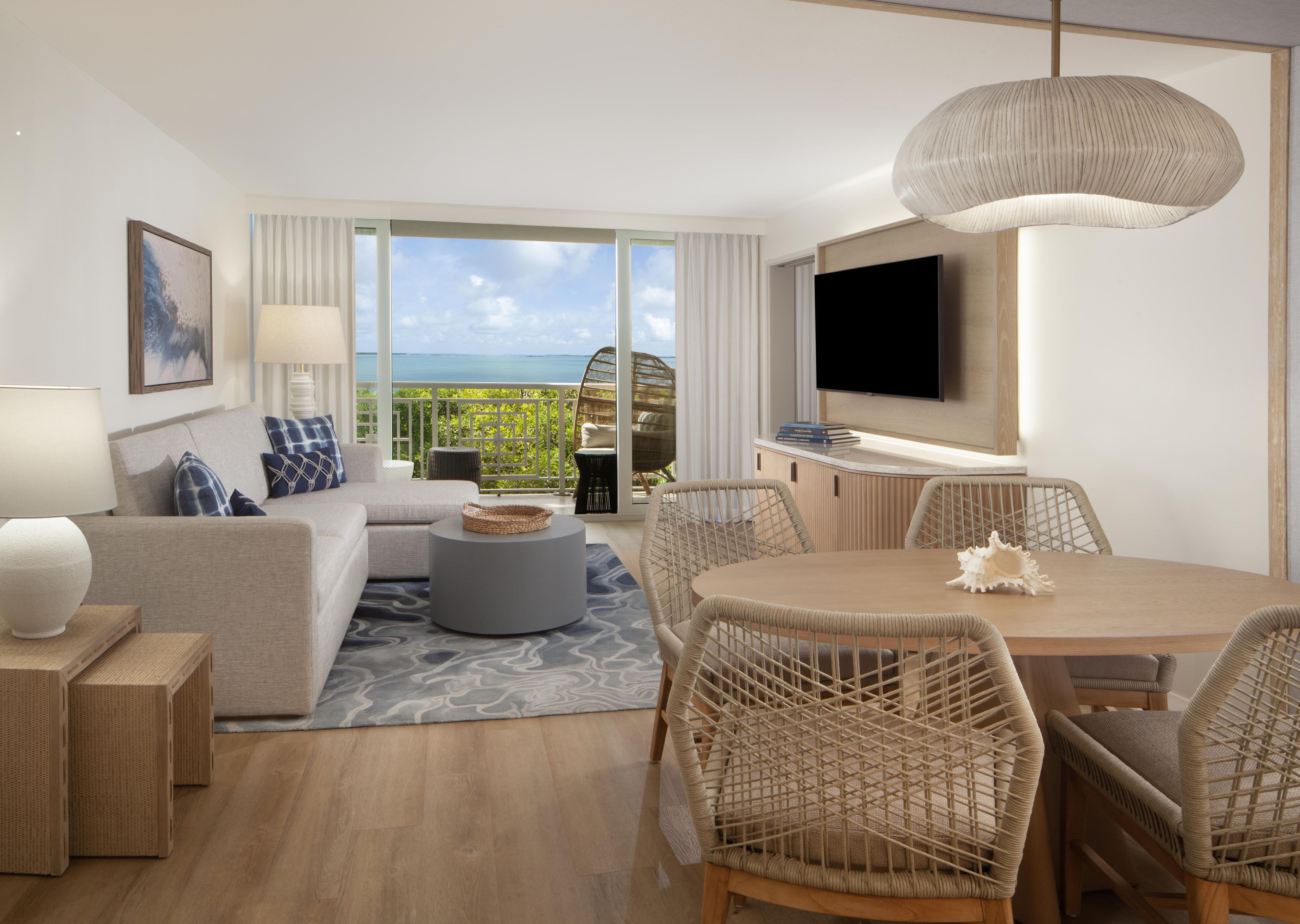 A modern hotel room with a sofa, dining table, chairs, a TV, and a balcony with a view.