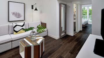 A modern living room with a sofa, a coffee table, and a large plant. There is a framed artwork on the wall and a door leading to a balcony.